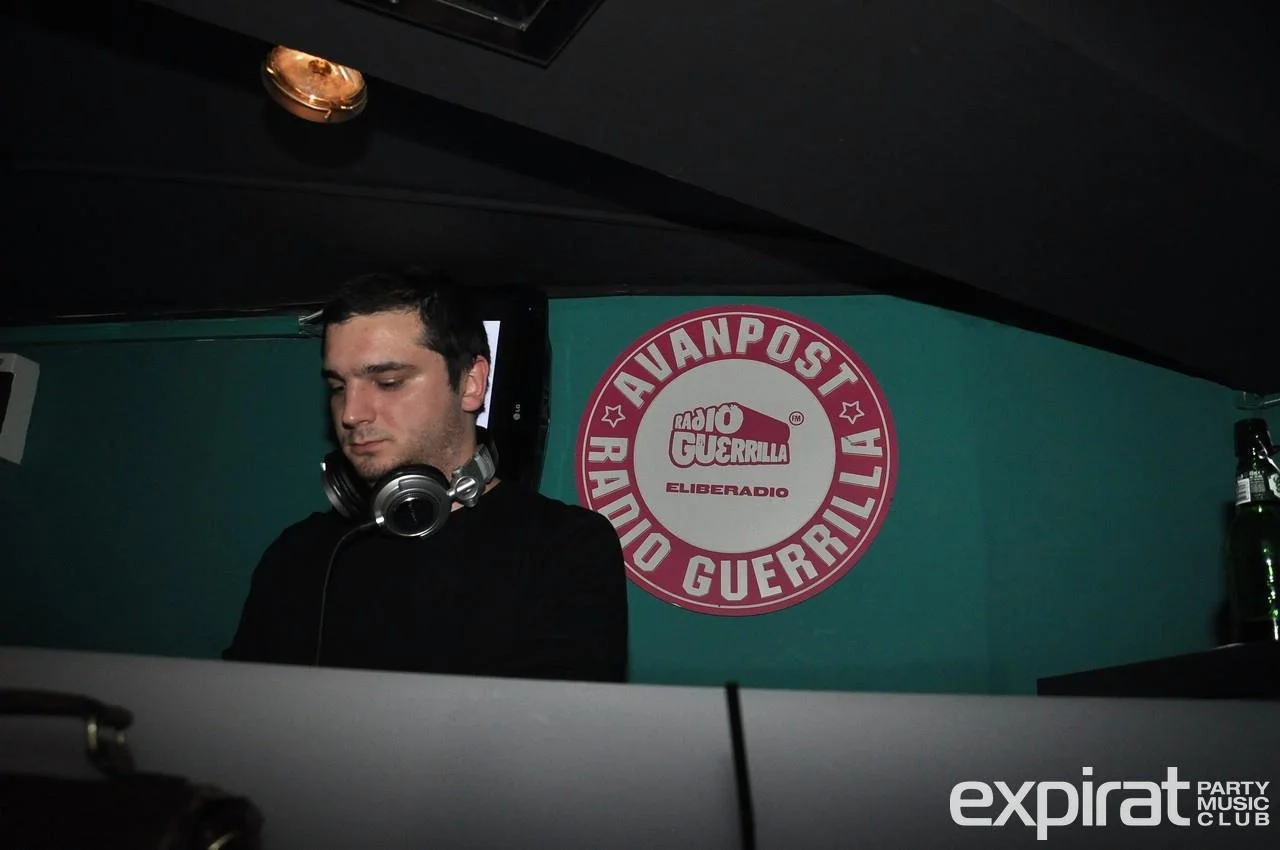 A DJ man with headphones around his neck is performing at a club, with a green wall behind him decorated with a circular logo that reads "Radio Guerrilla" and "Avanpost."