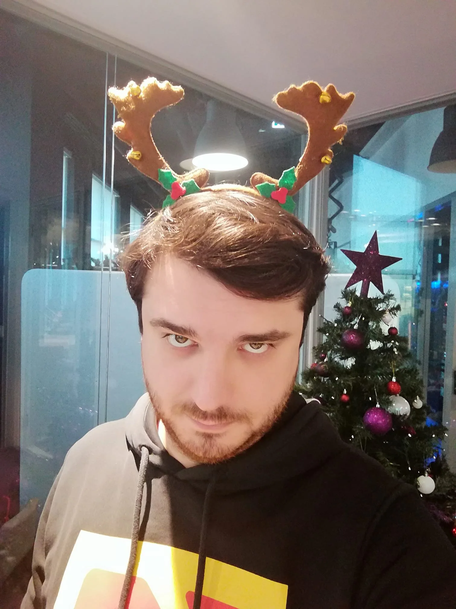 A man taking a selfie wearing reindeer antlers headband with Christmas decorations, standing in front of a decorated Christmas tree.