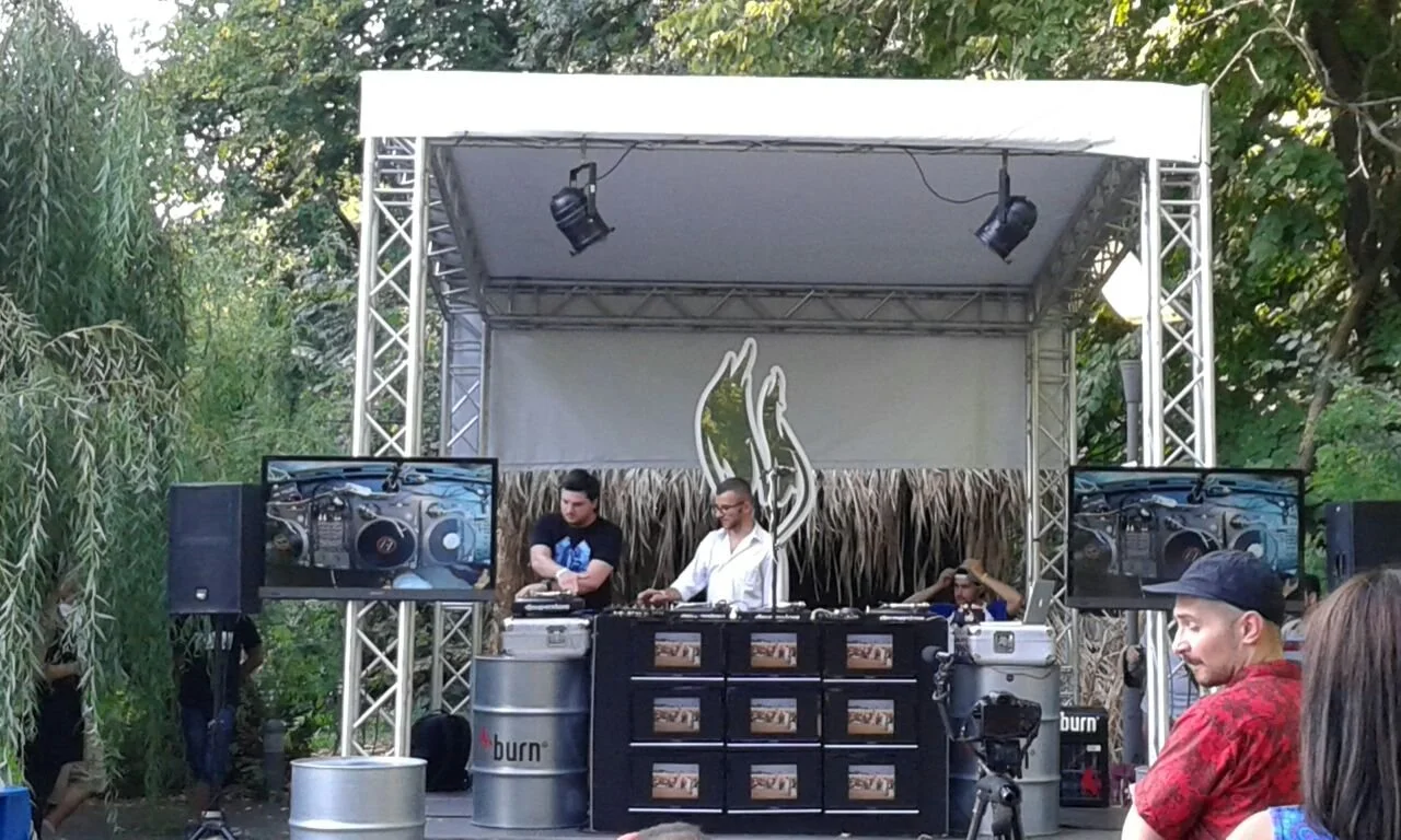 A DJ performing on a small outdoor stage with two large monitors, an audience, and lush greenery in the background.