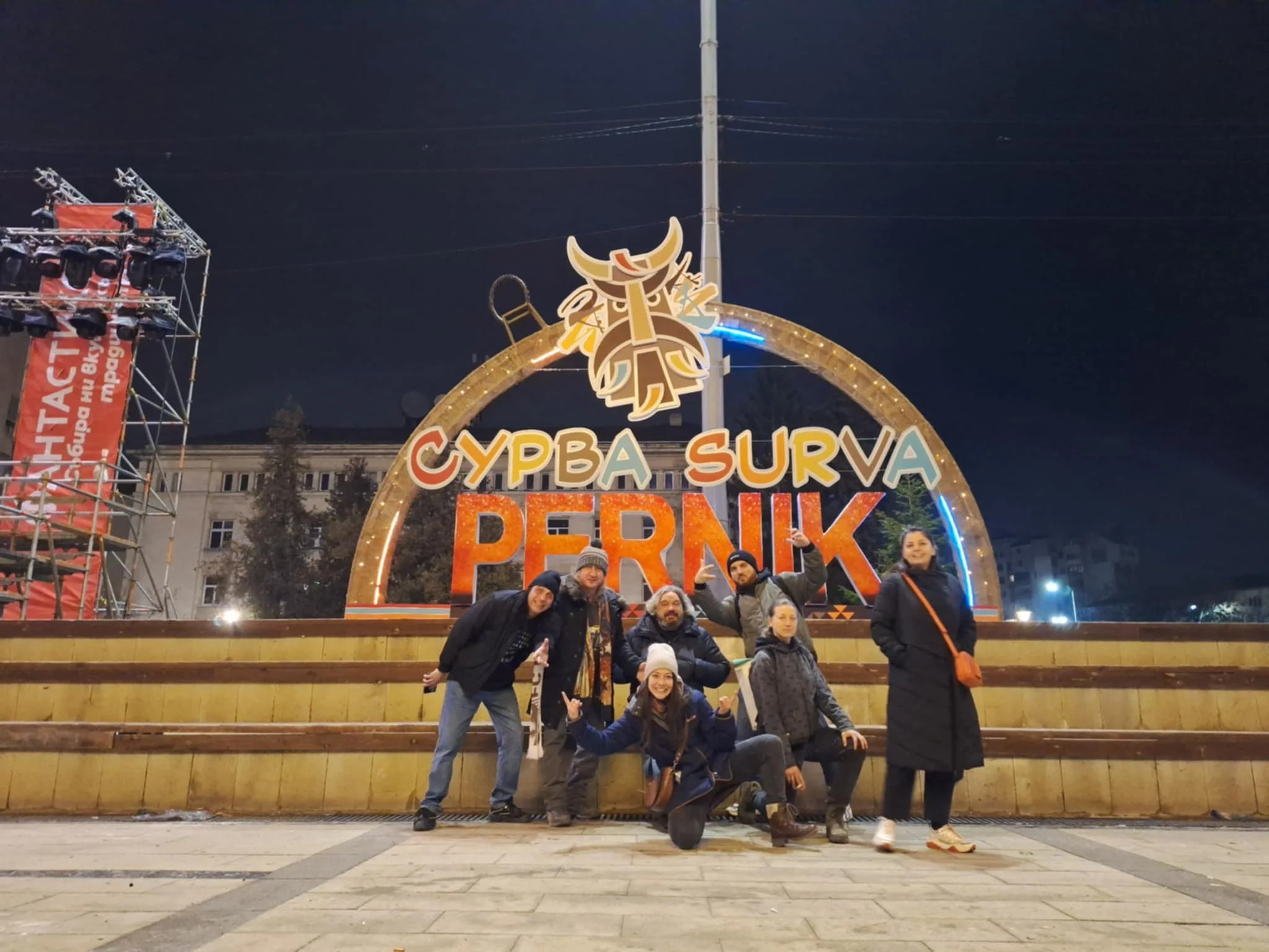 Group of six people posing in front of a decorative sign at night in Pernik, Bulgaria, with a wooden platform and buildings in the background.