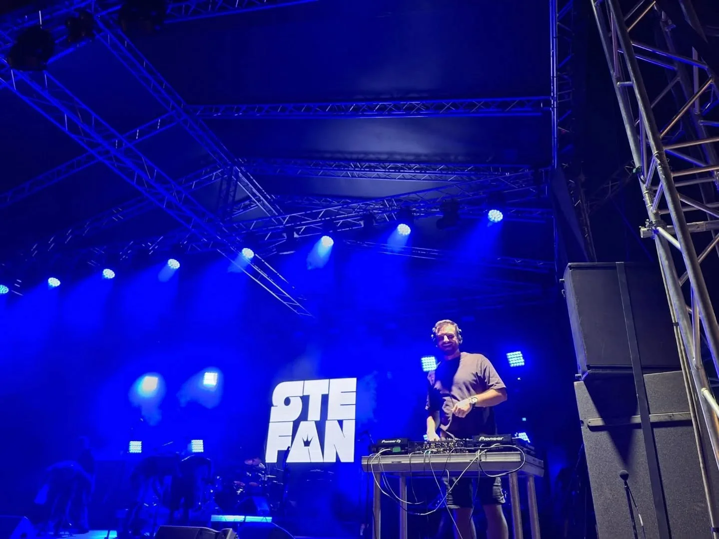 A DJ stands behind turntables on stage with blue stage lights, large screen displaying "STE FAN," and metal trusses overhead at an event or concert.