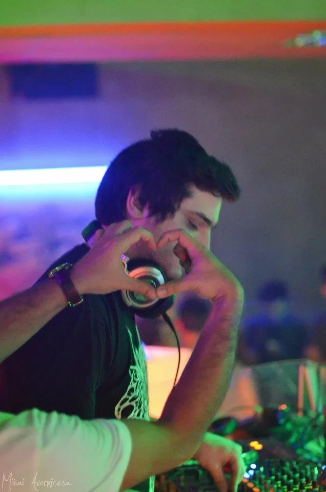 A young man with headphones around his neck making a heart gesture with his hands at a nightclub or music event with colorful lighting.