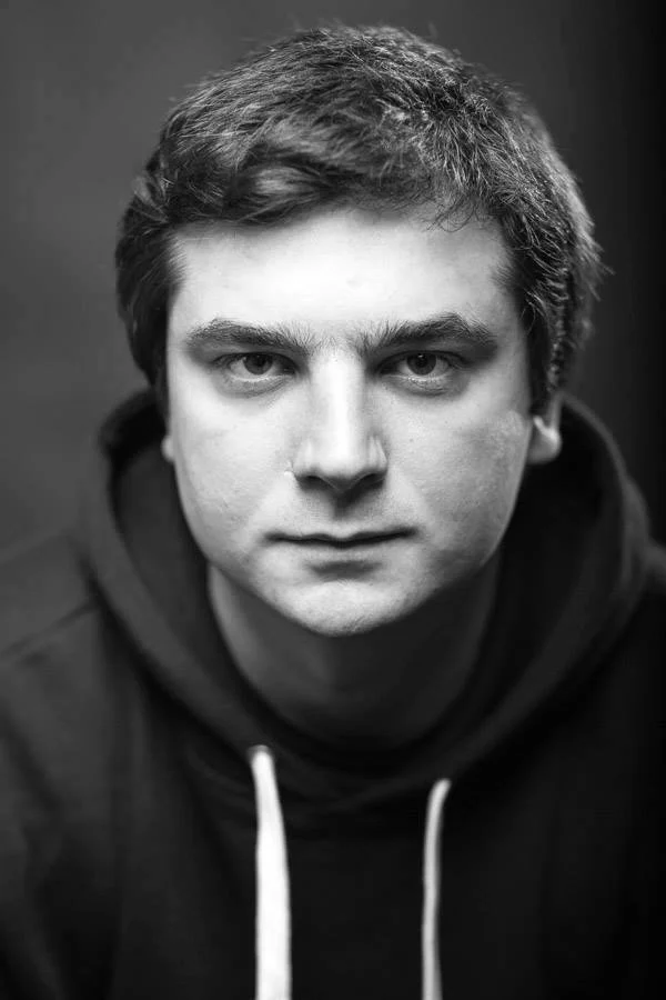 Black and white portrait of a young man with short hair, wearing a hoodie, looking directly at the camera with a serious expression.