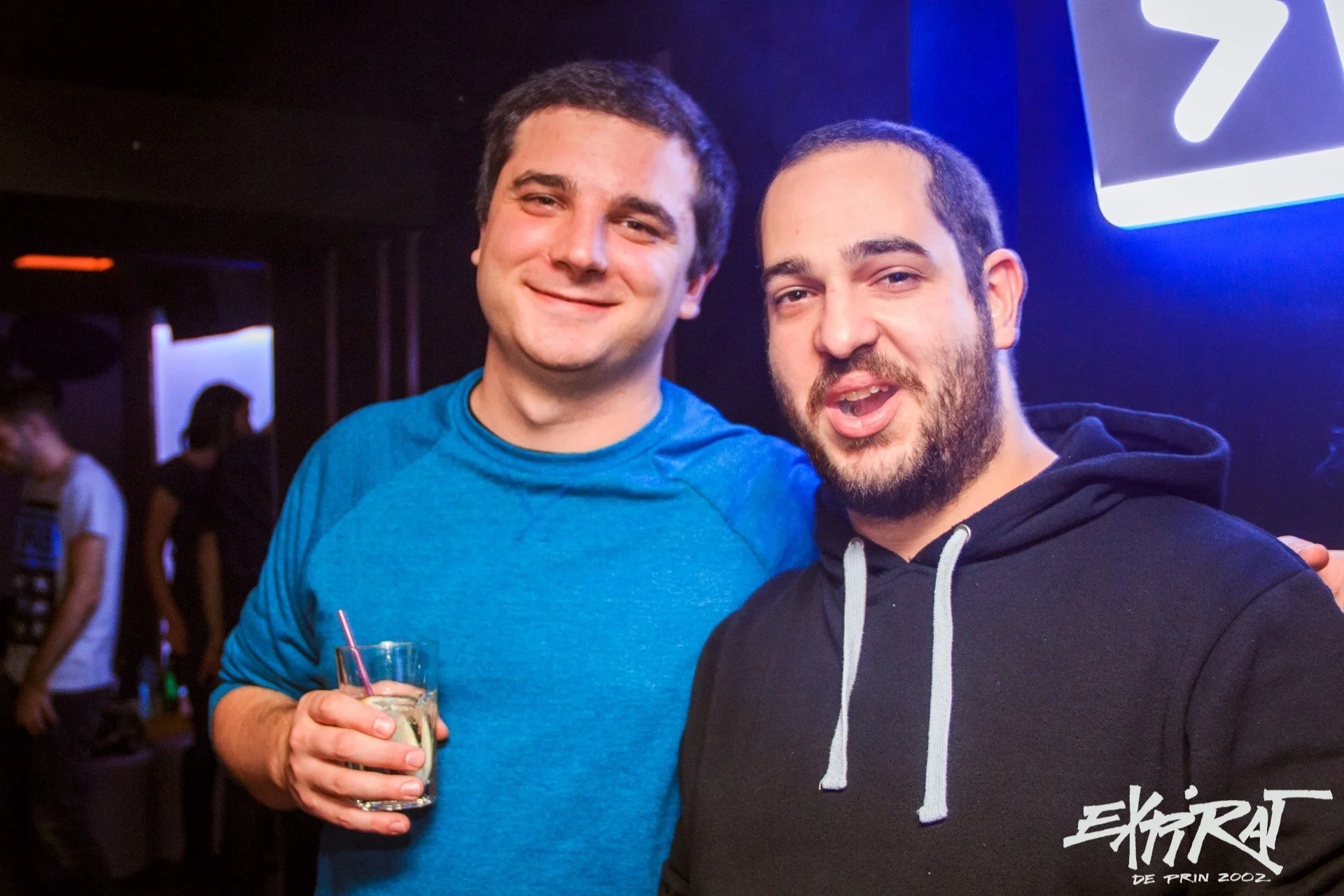 Two men smiling and posing for a photo at a social event, one holding a glass with a drink, in a dimly lit venue with other people in the background.