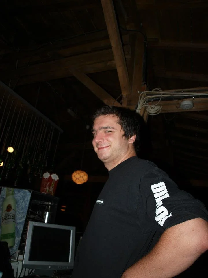 A man with dark hair and a slight smile wearing a black shirt with white text on the sleeve, standing inside a dimly lit room with a wooden ceiling and various objects, including a monitor and bottles, in the background.