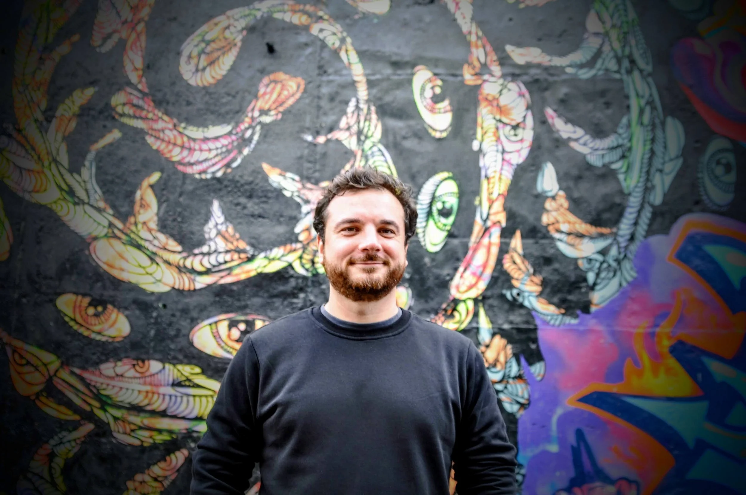 A man with a beard in a black shirt standing in front of a colorful graffiti mural on a wall.