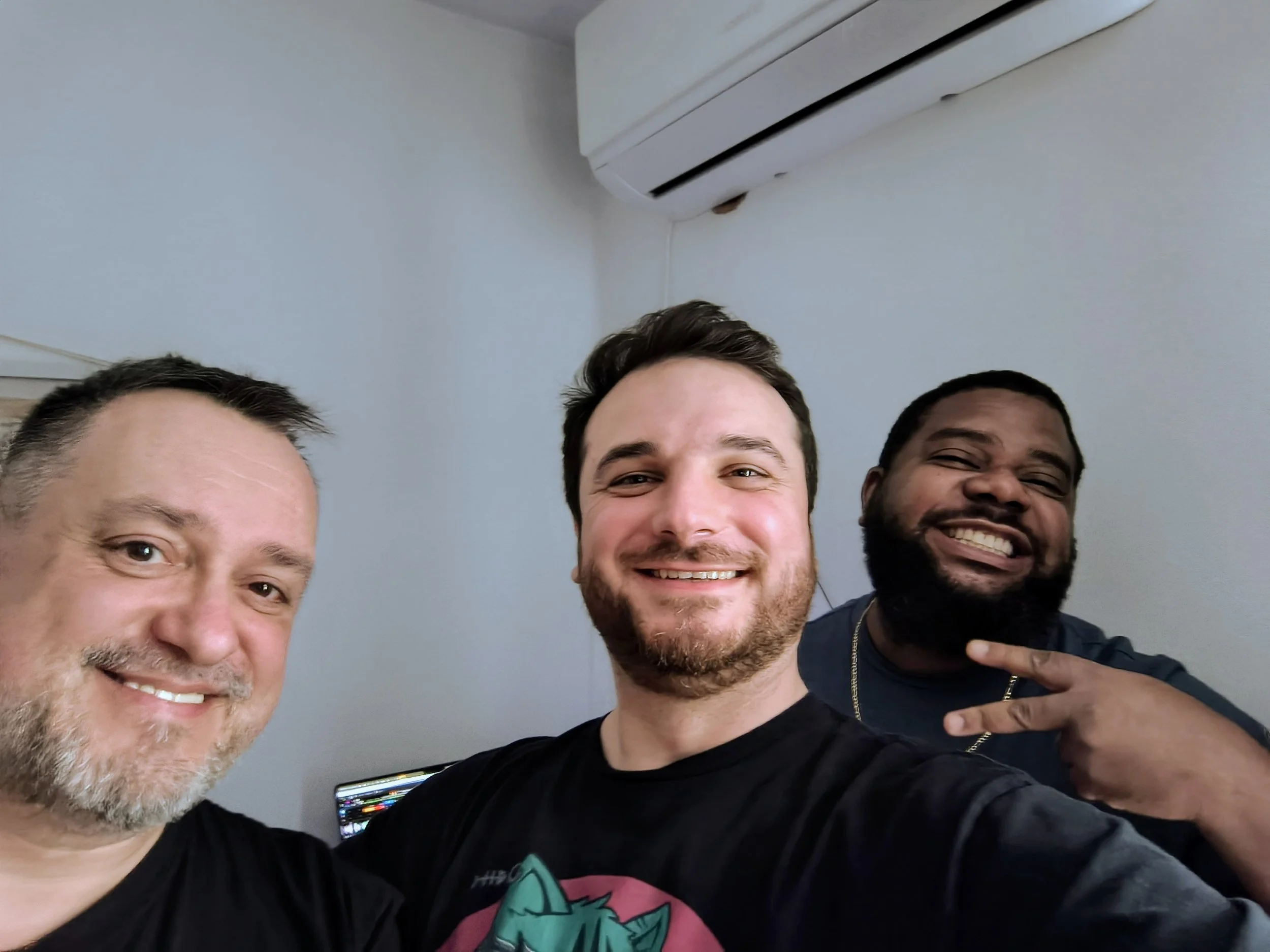 Three men smiling and posing for a selfie in an indoor setting, with an air conditioning unit visible on the wall behind them.