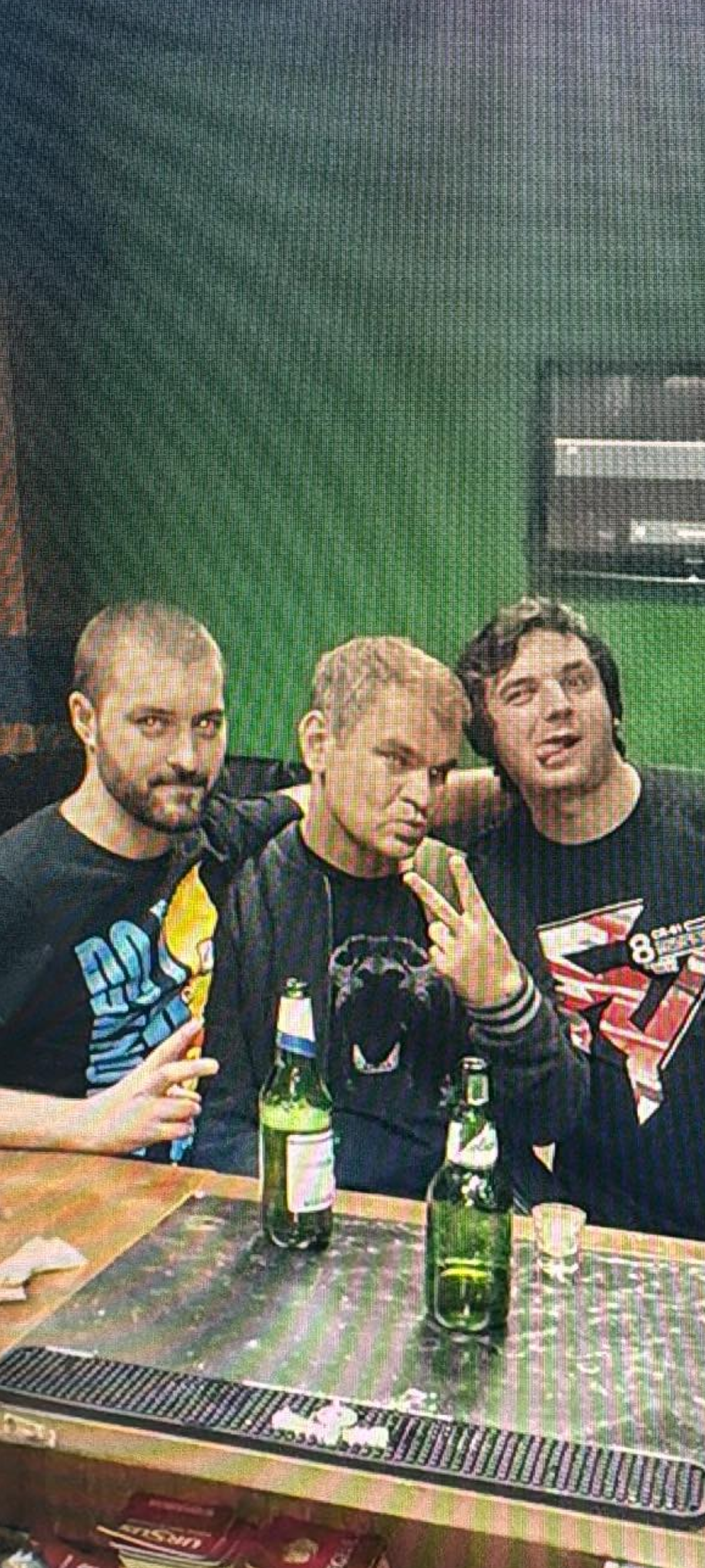 Three men sitting together at a table with beer bottles, in front of a green wall. They are posing for the photo, with the man in the middle making a peace sign.