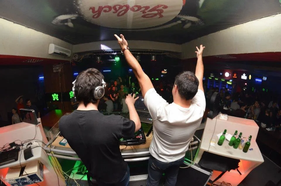Two DJs perform at a nightclub with a crowd dancing in the background. The DJ on the left wears headphones, and the DJ on the right raises his hands. Several green beer bottles are on the DJ booth.