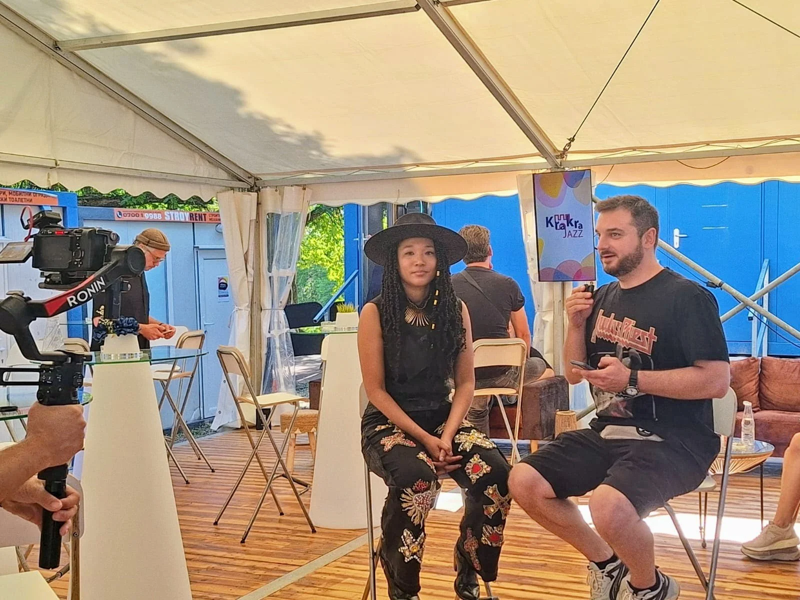 Two people are seated on chairs inside a tent, one is a woman with long dark hair wearing a black hat and patterned pants, the other is a man with short hair and a beard wearing a black t-shirt and shorts. There is filming equipment and other people 