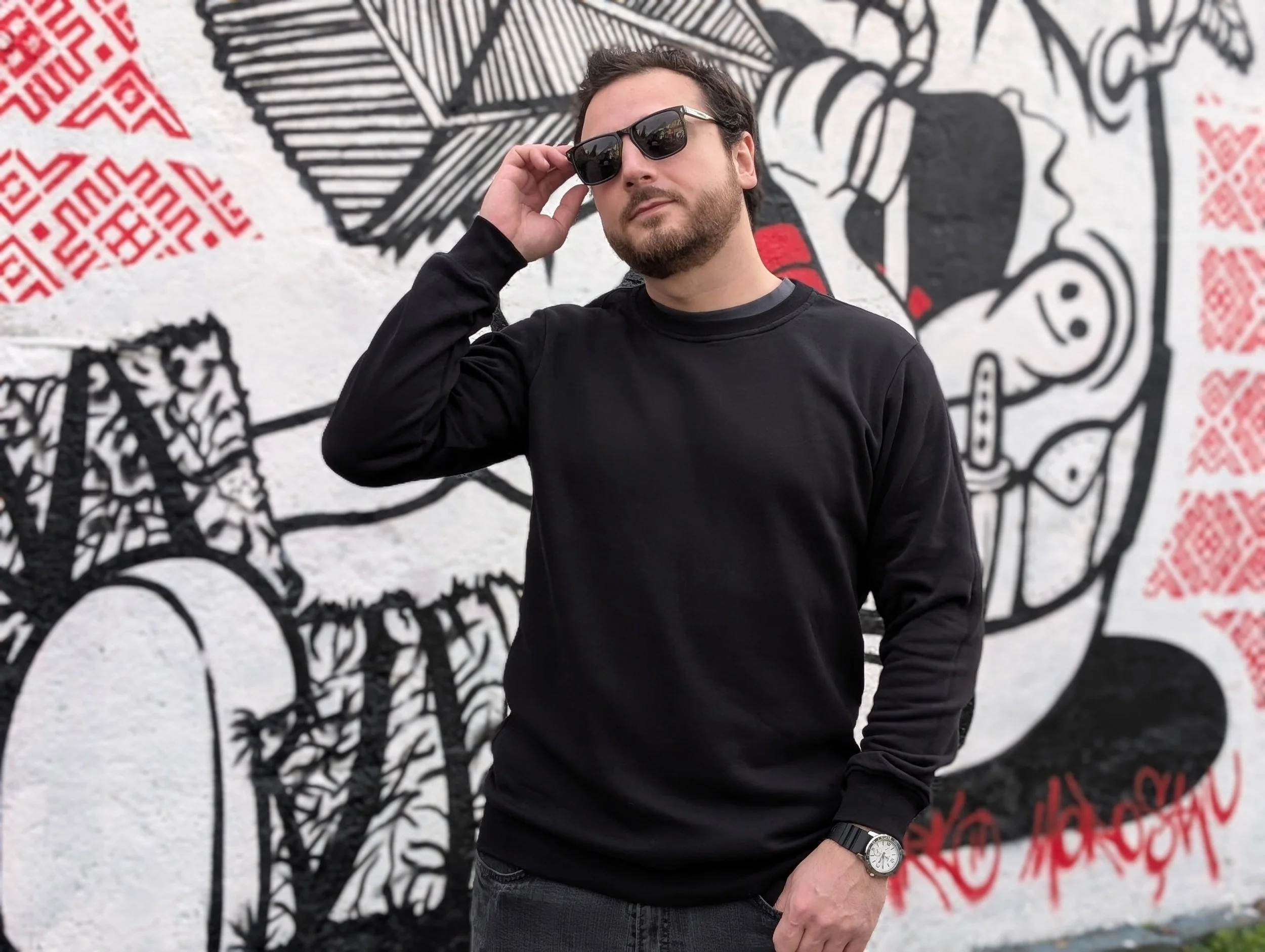 A man with sunglasses and a watch standing in front of a graffiti wall featuring black, red, and white designs.