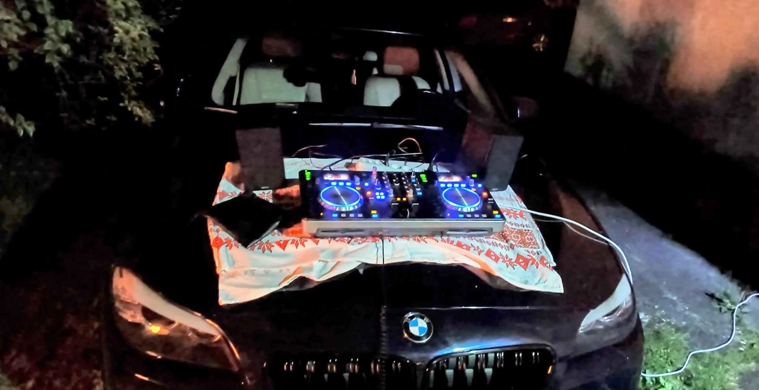 Nighttime scene of a DJ setup with a mixer and turntables on the hood of a black BMW car, which is parked outdoors near bushes, with a cloth underneath the equipment.