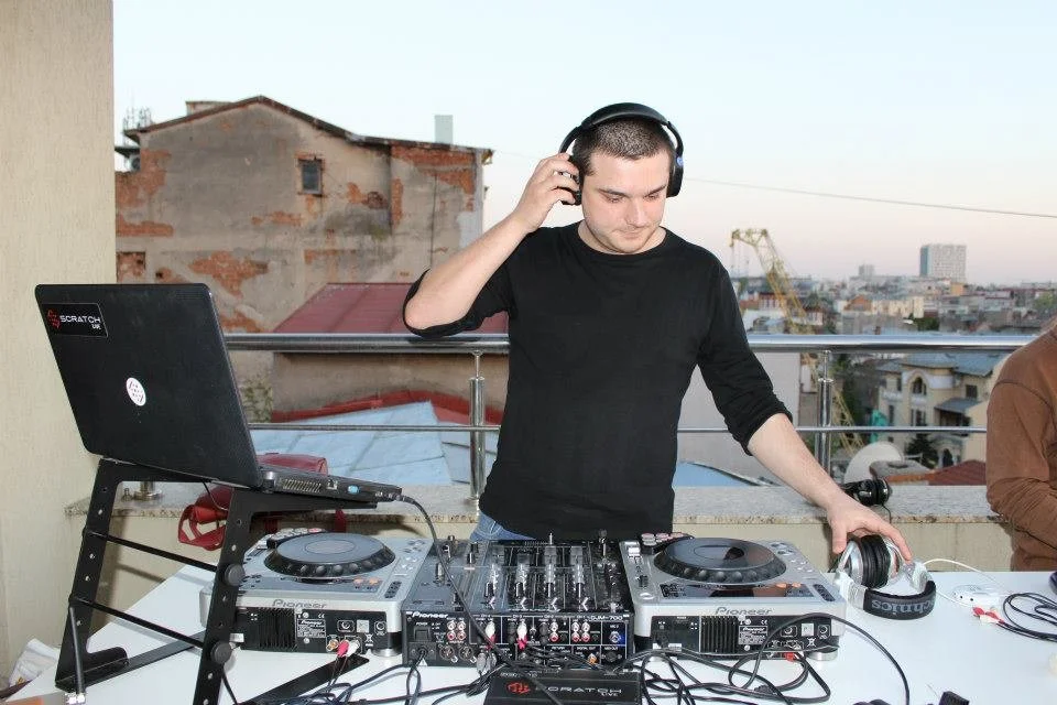 A DJ wearing headphones at a rooftop party with turntables, a laptop, and mixing equipment, with an urban cityscape in the background.