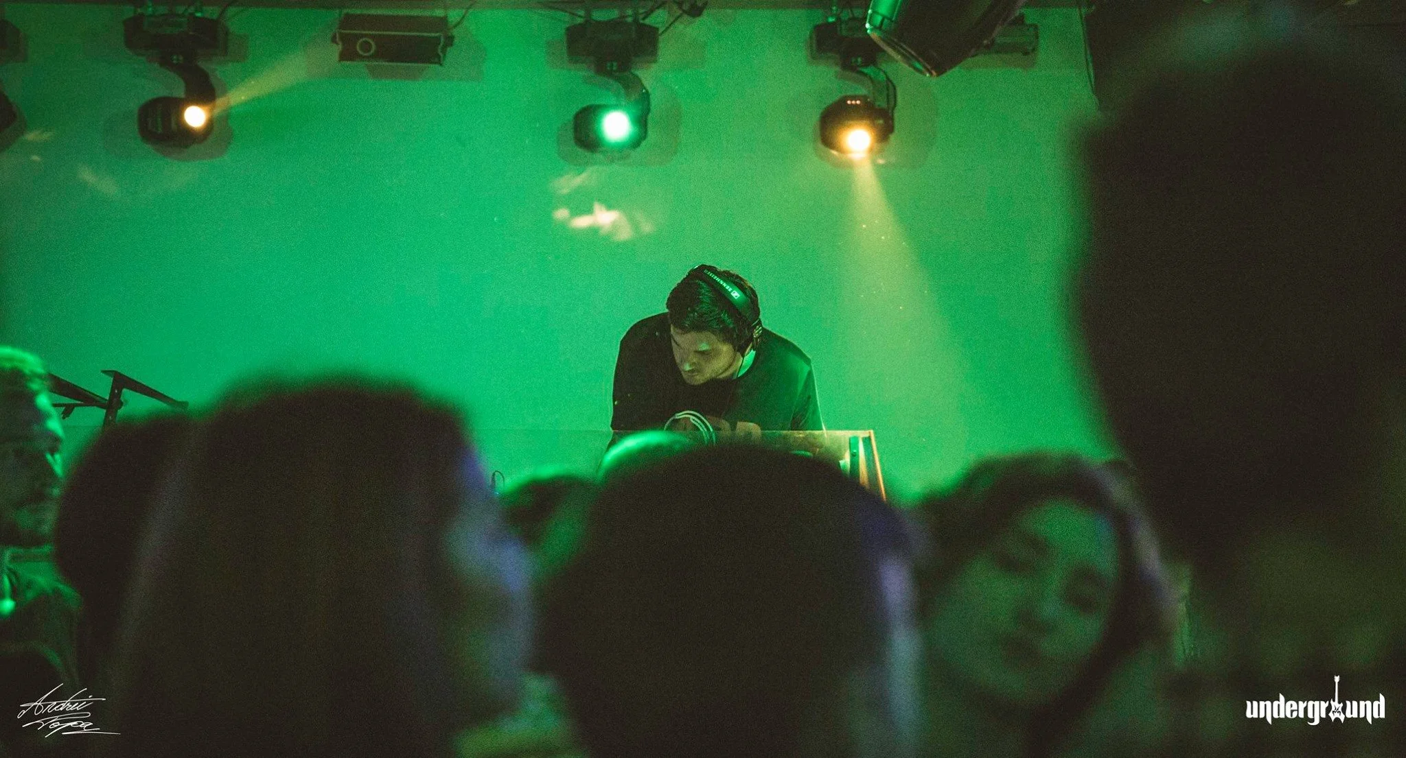 A DJ performing at a nightclub with green lighting, wearing headphones and standing behind a DJ booth, with an audience in the foreground.