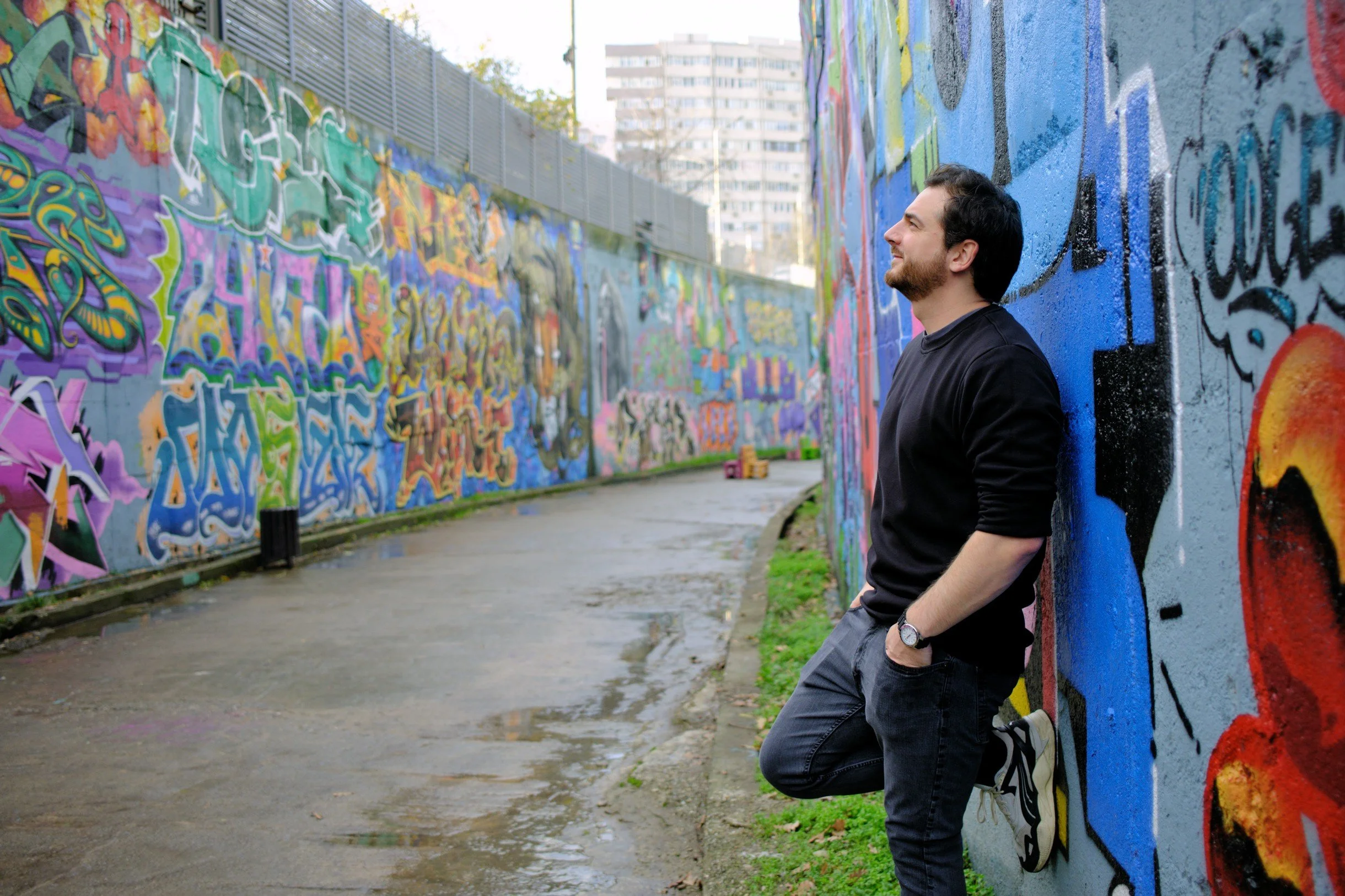 A man with dark hair and a beard leaning against a graffiti-covered wall, with a pathway and buildings in the background.