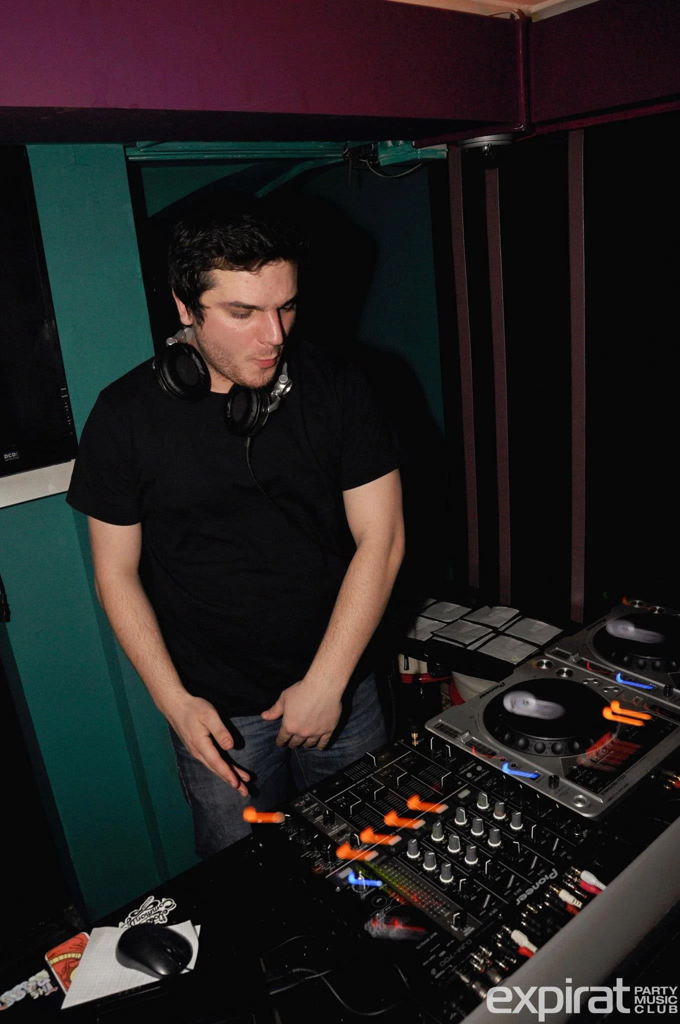 A DJ standing in front of turntables and audio equipment, wearing a black t-shirt and headphones around his neck, in a nightclub or music venue.