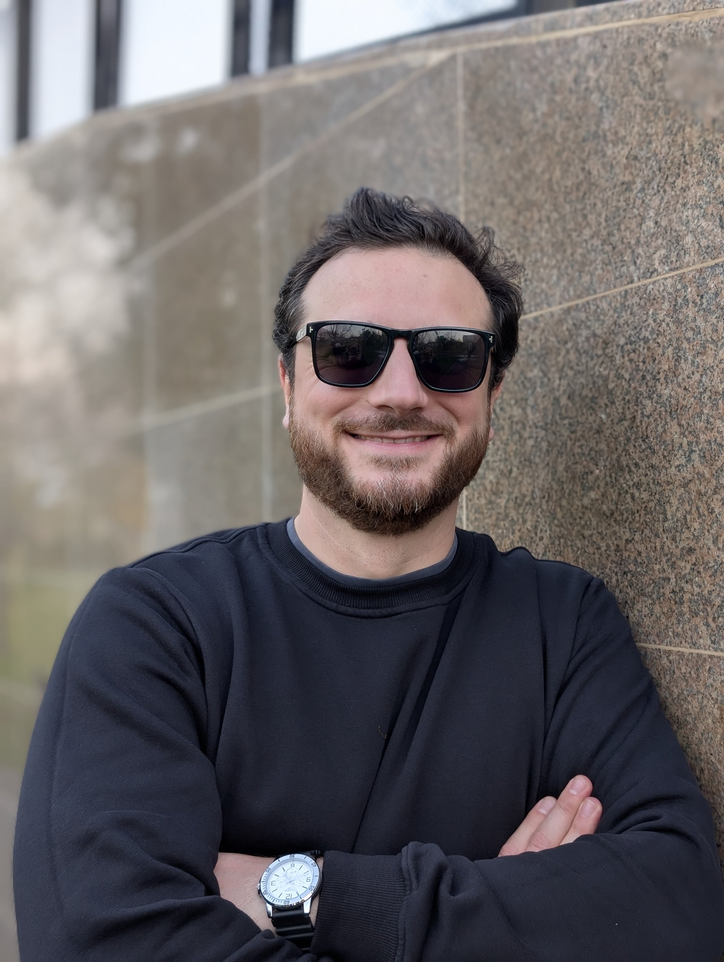 A man with dark hair and a beard wearing sunglasses and a black long-sleeve shirt, standing outdoors with arms crossed in front of a stone wall.