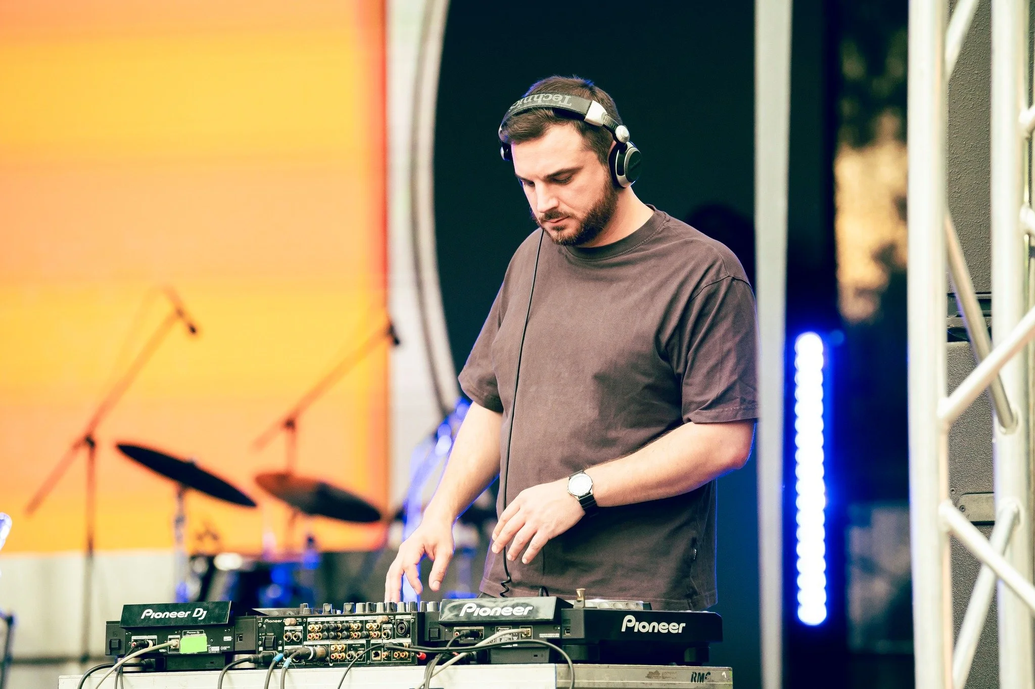 A DJ wearing headphones and a watch while operating DJ equipment with microphones and drums in the background at an outdoor event.