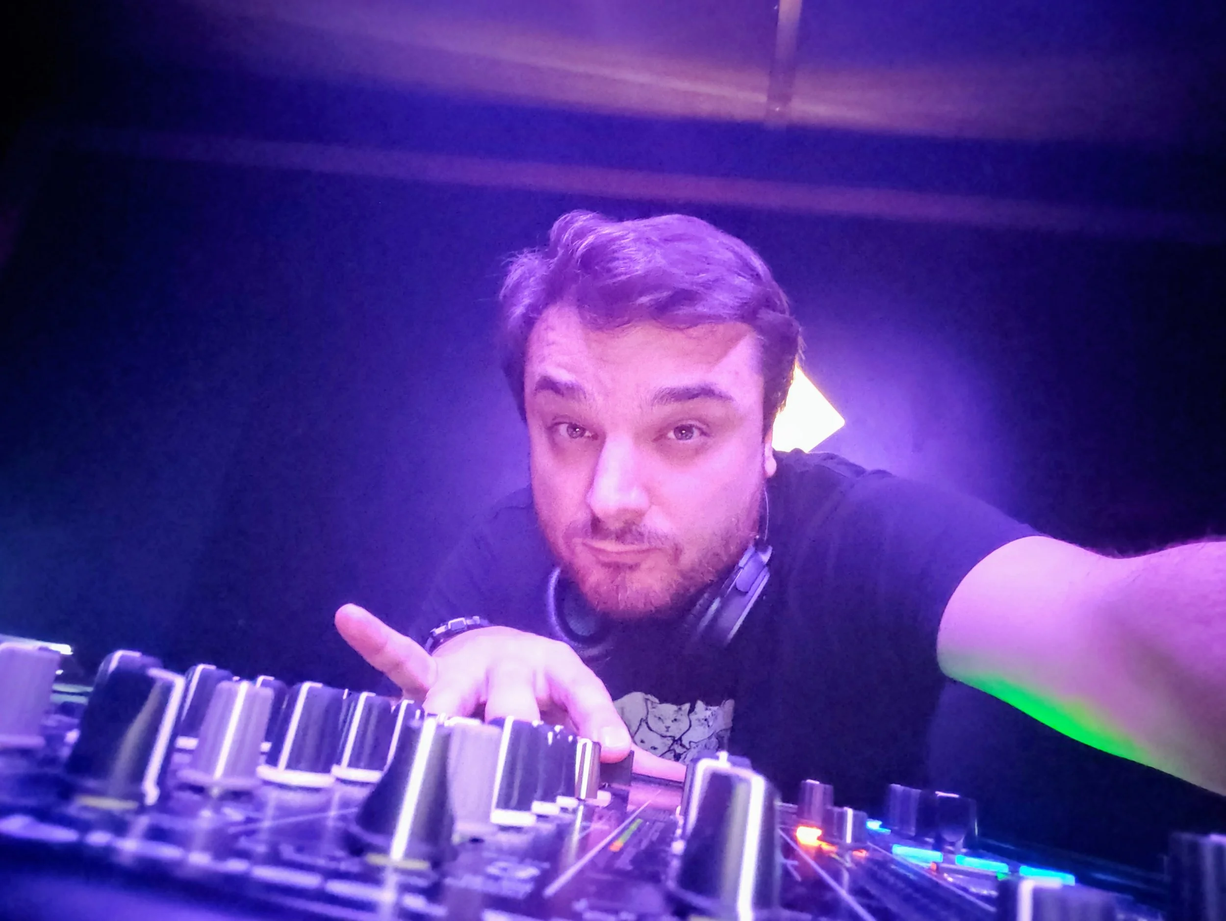 A young man with shoulder-length brown hair, wearing a black T-shirt and headphones around his neck, is taking a selfie in front of a DJ mixer under purple and green lighting.