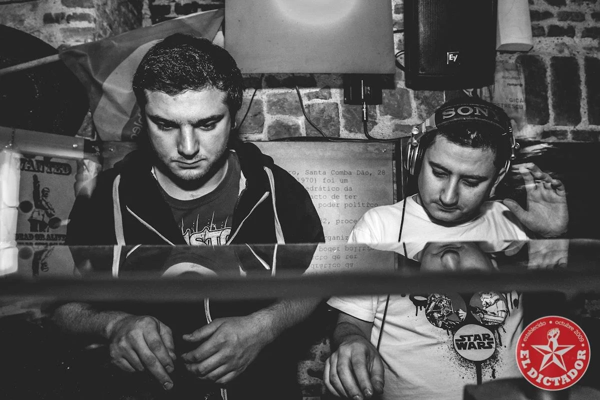 Two young men are working as DJs in a music venue, one wearing headphones and both focused on their equipment, with a brick wall in the background.