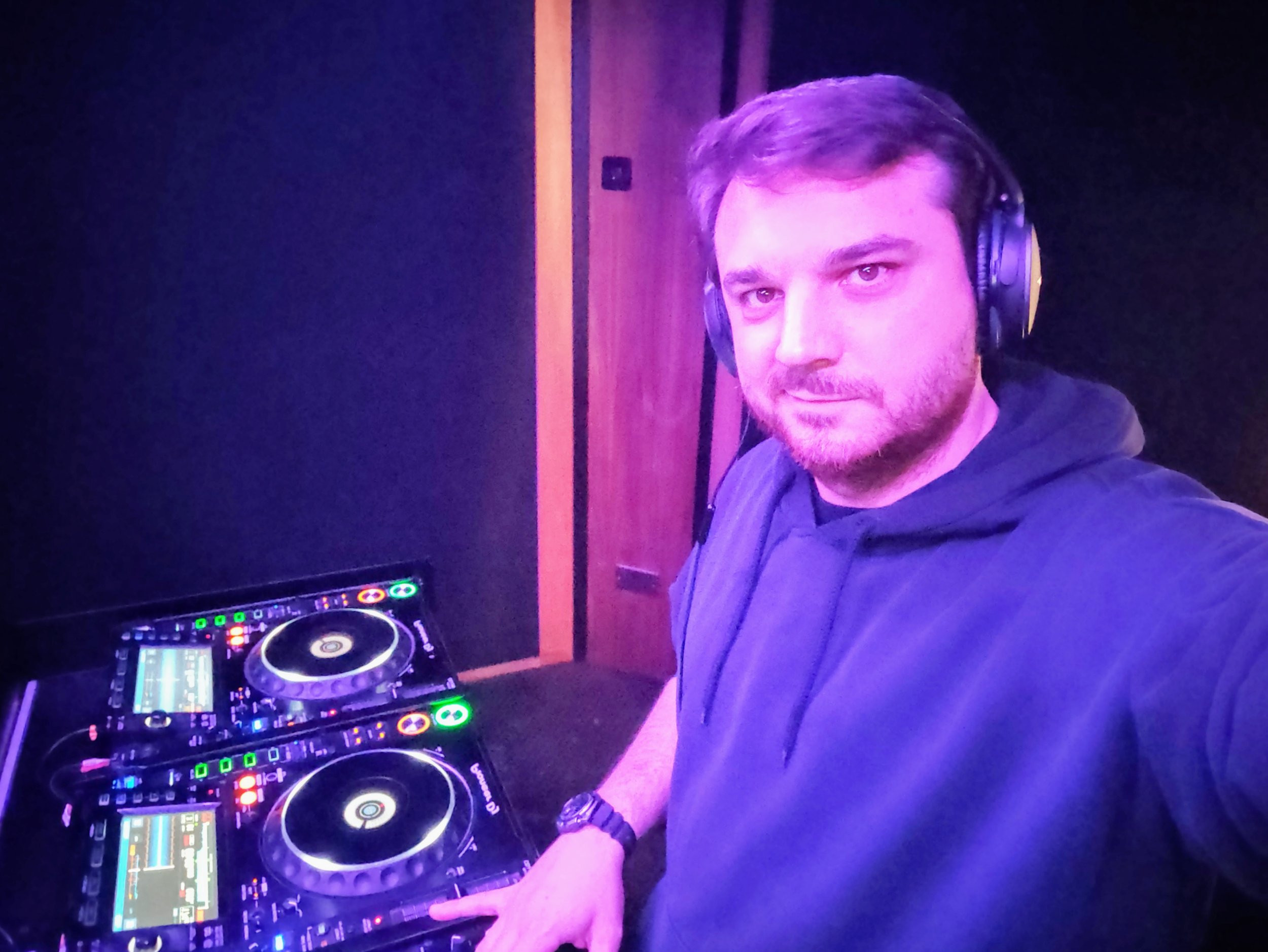 A man with headphones standing in front of DJ equipment, including turntables with bright lights and screens, in a dimly lit room with purple and blue lighting.
