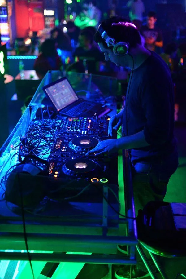 A DJ wearing headphones operates turntables and a mixer at a nightclub with colorful LED lighting and blurred clubgoers in the background.
