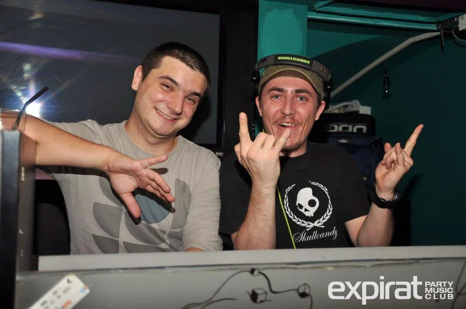 Two men are smiling and making peace signs at a party or nightclub, with one wearing headphones and the other wearing a team shirt.