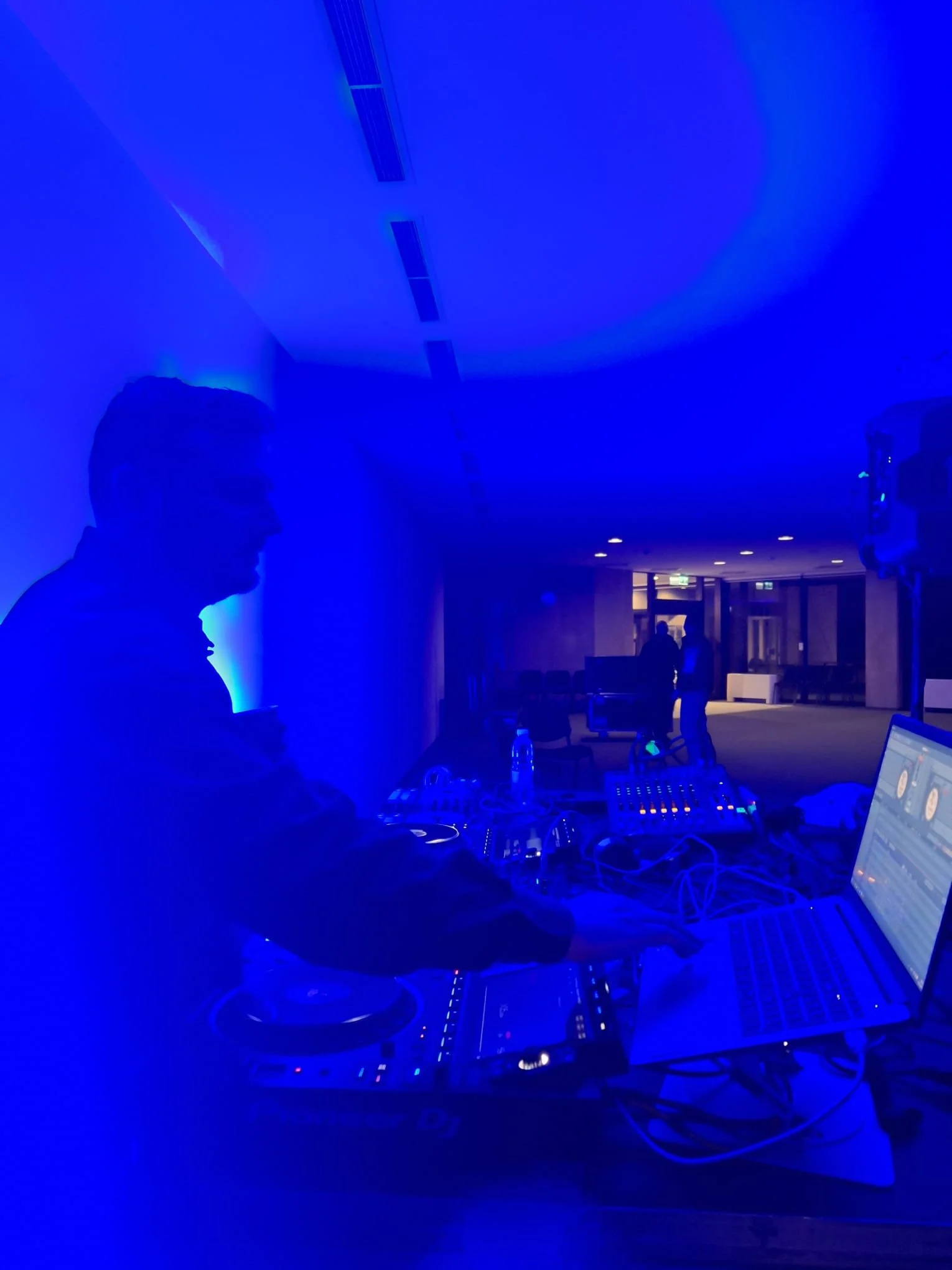 A DJ performing at an event in a dimly lit room bathed in blue light, with equipment, a laptop, and people in the background.