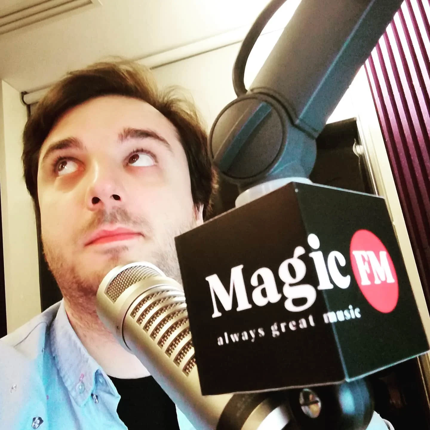 A man with light skin and brown hair sitting at a radio microphone with a "Magic FM" sticker, looking upward with a thinking expression.
