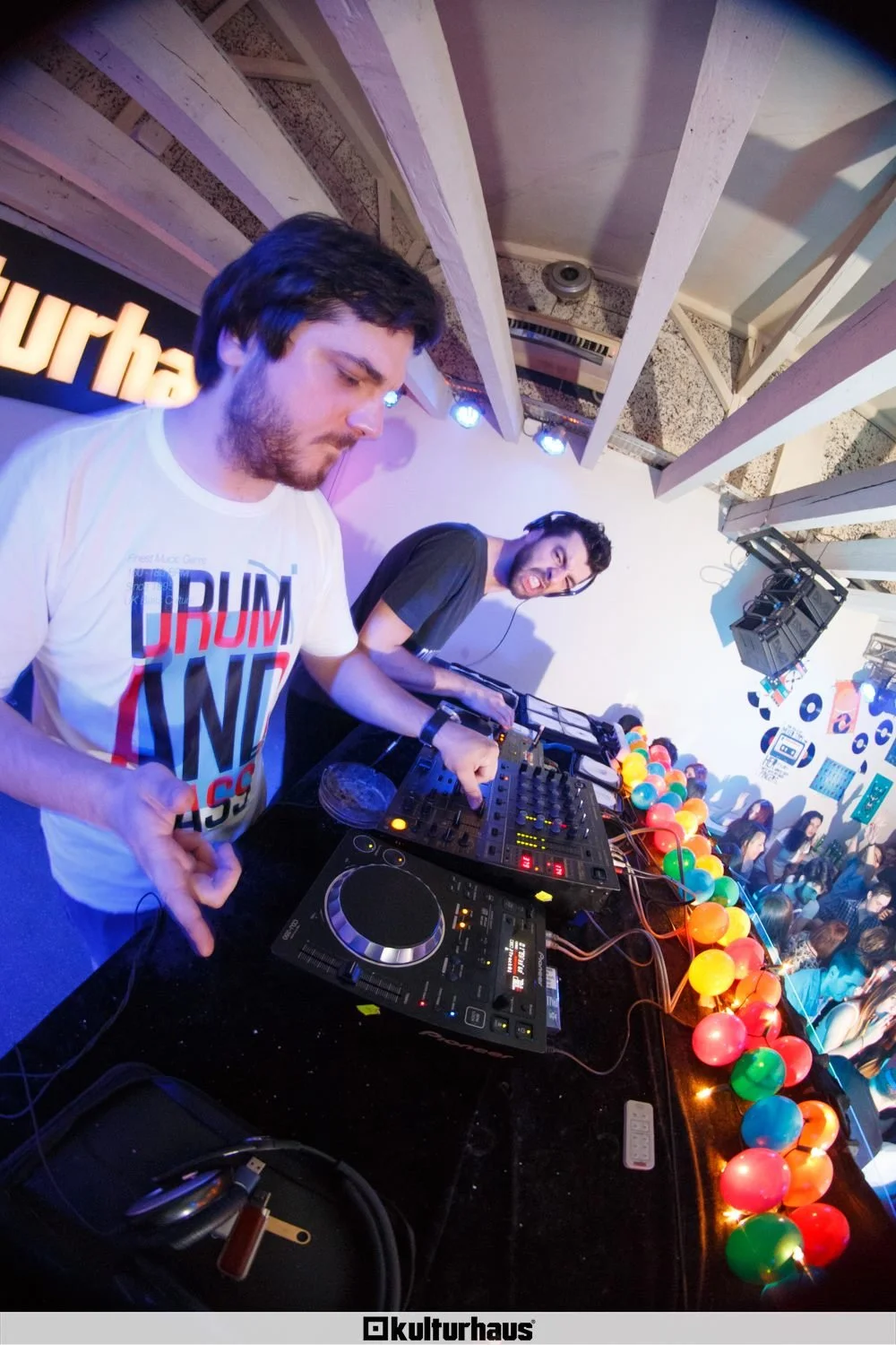 Two male DJs perform at a nightclub, with one wearing a white t-shirt with colorful text and the other wearing a black t-shirt. They are using DJ equipment on a black table decorated with colorful string lights. Behind them, there are rotating colore