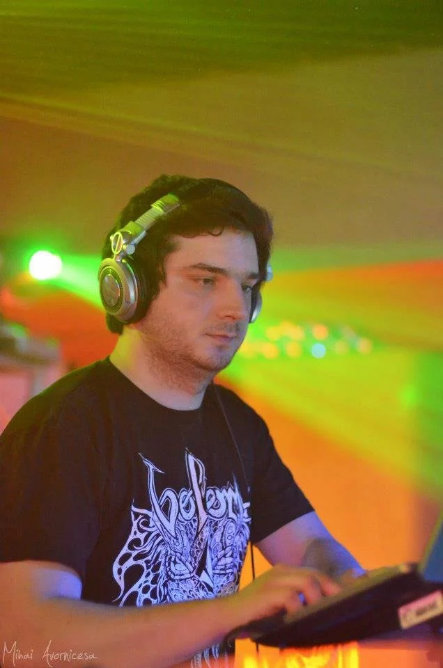 A young man with dark hair and headphones in a dimly lit room, working on a laptop with colorful green and orange lighting background.