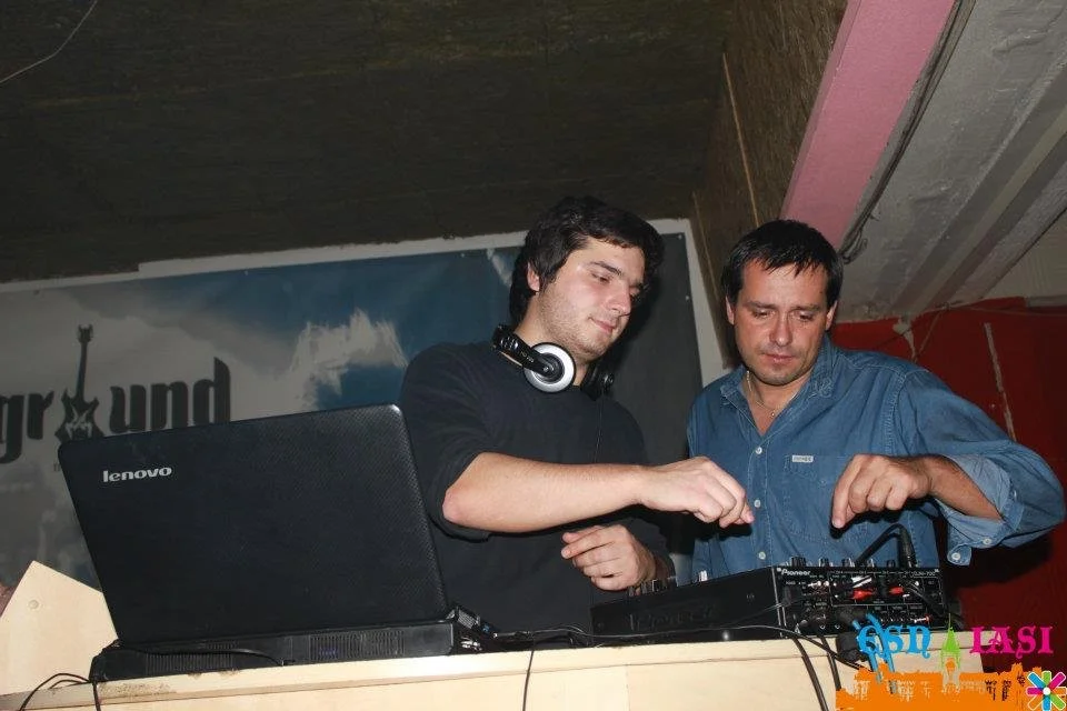 Two men working with DJ equipment, one wearing headphones, in a dimly lit room with a poster in the background.
