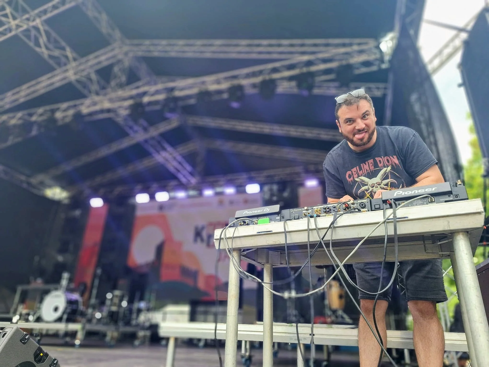 DJ with a beard and sunglasses on his head, smiling and sticking out his tongue, working at a DJ booth on an outdoor stage with metal scaffolding, with a large colorful banner and stage equipment in the background.
