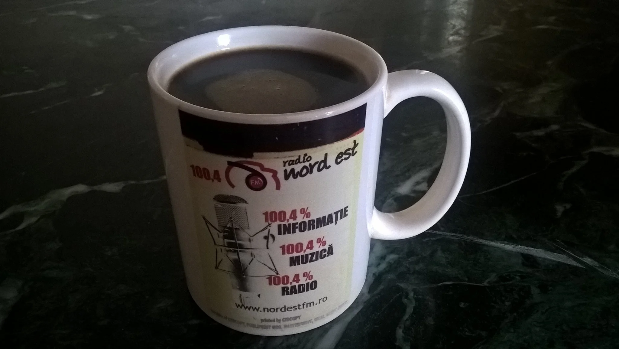 White mug filled with black coffee sitting on a dark marble surface, with a logo and text for Radio Nord Est