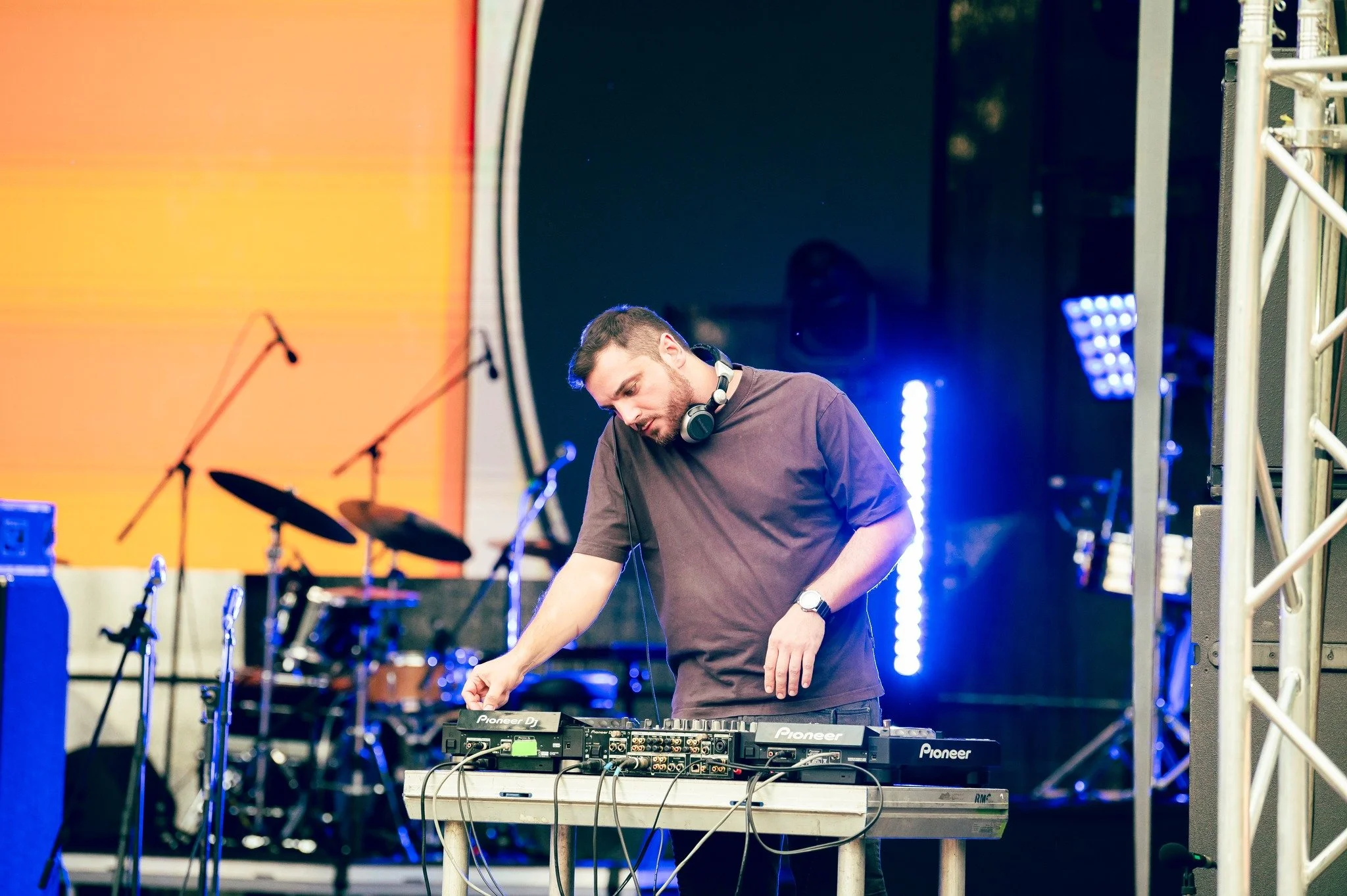 A DJ wearing headphones standing at a DJ mixer with a microphone and drums in the background on stage, with colorful stage lighting.