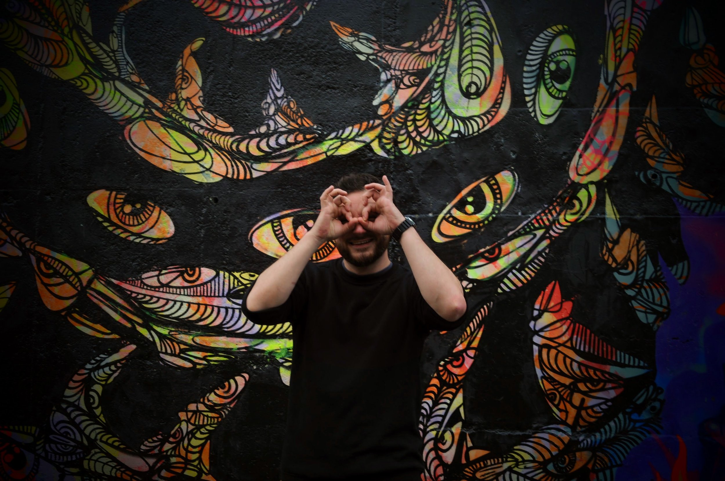 A smiling man making heart shapes with his hands around his eyes, standing in front of a vibrant, abstract mural with swirling, colorful patterns on a black background.