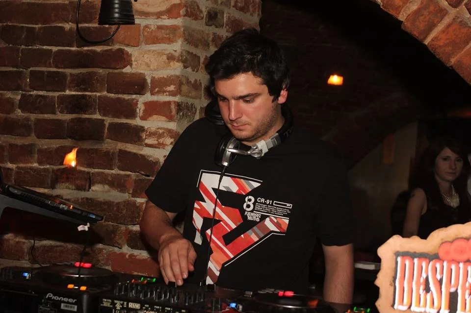 A young male DJ with dark hair, wearing a black graphic T-shirt and headphones around his neck, is focused on mixing music at a brick wall venue. A woman with long dark hair and a black dress is visible in the background.