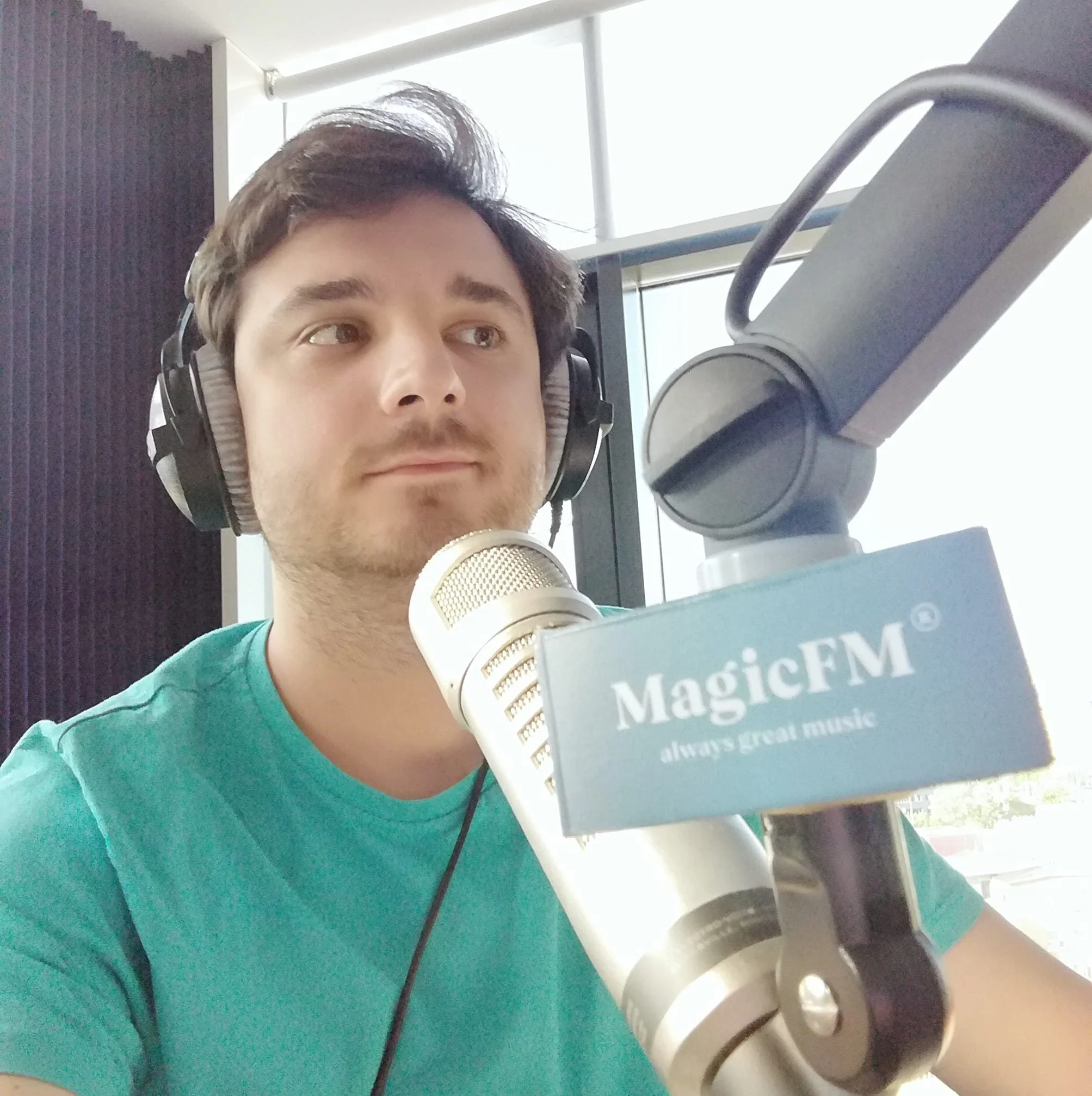 A young man wearing headphones sits in front of a microphone labeled MagicFM, with the tagline always great music, in a radio studio.