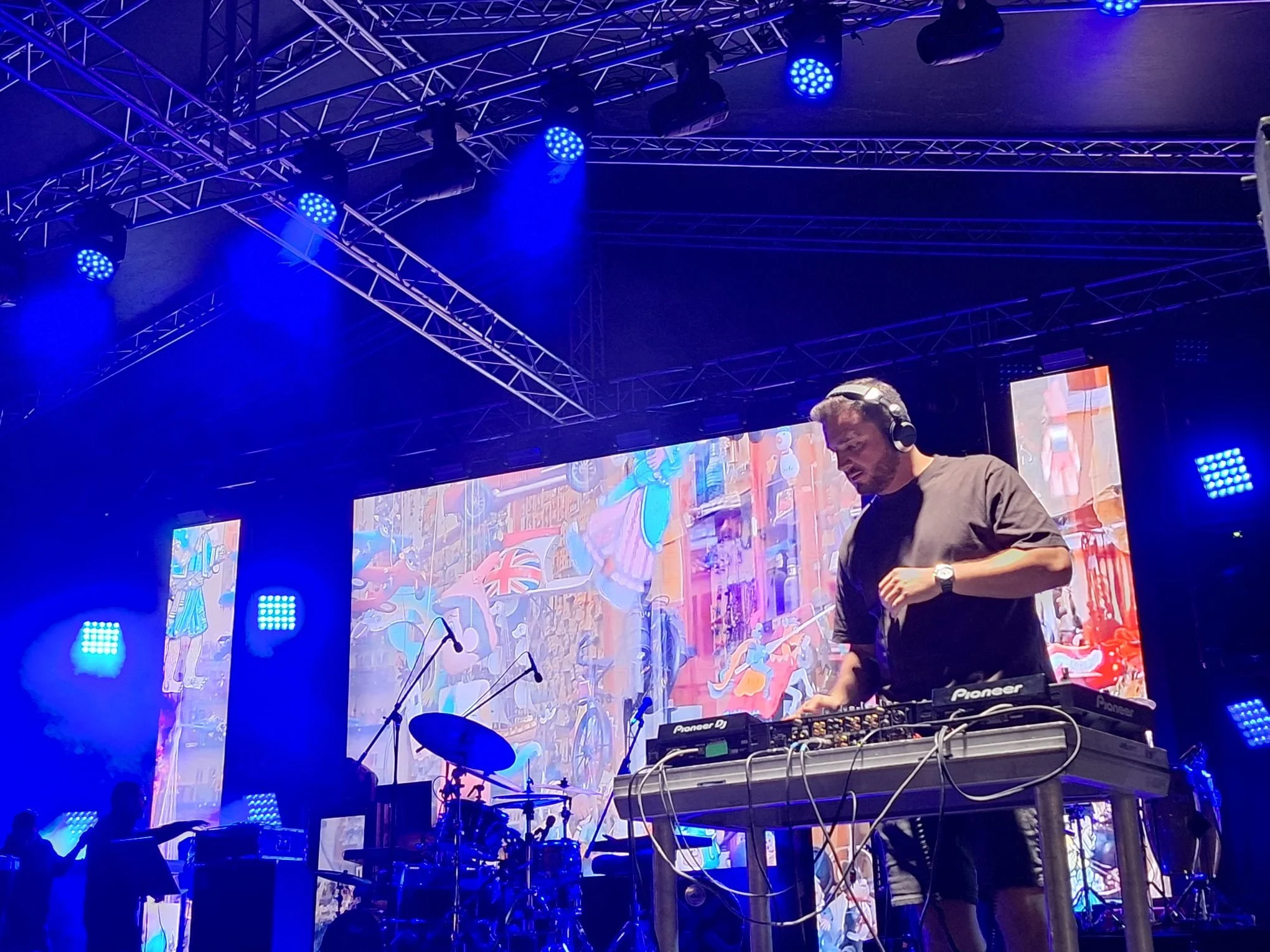 A DJ wearing headphones operates a Pioneer DJ console on a stage with colorful digital screens and blue stage lights, performing at a concert.