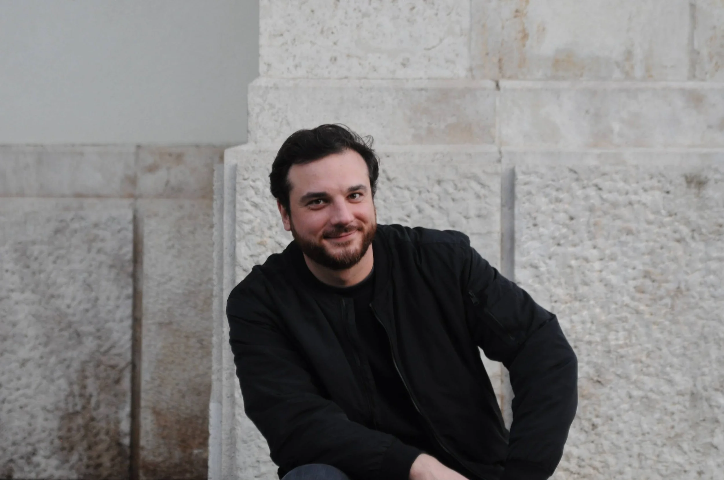 A man with dark hair and a beard wearing a black jacket, sitting on the ground with a stone wall in the background, smiling at the camera.