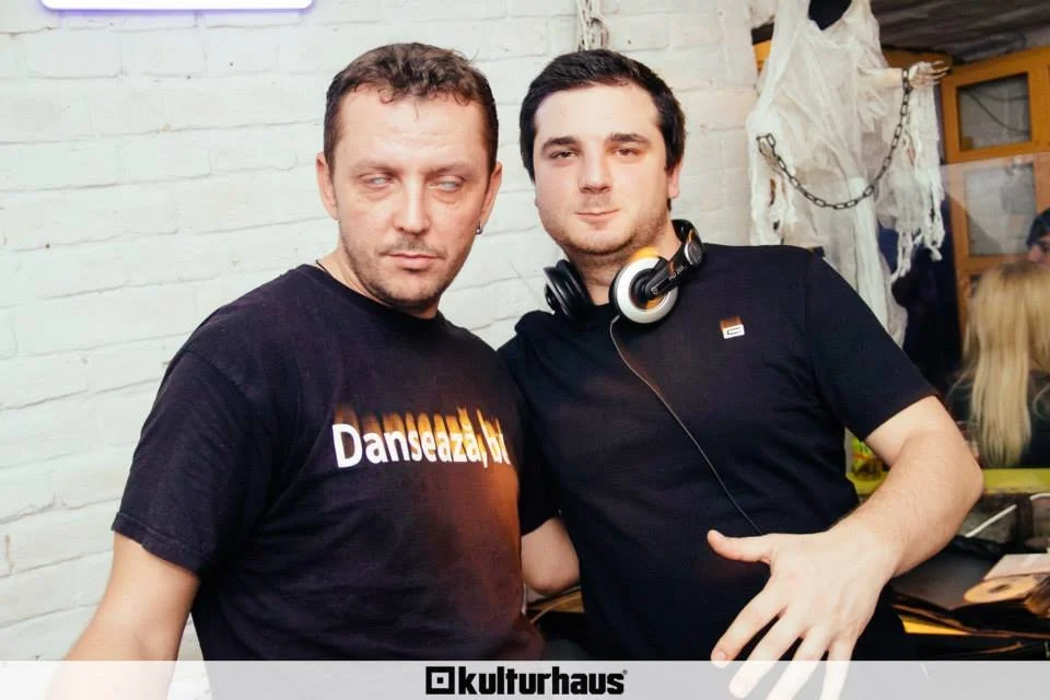 Two young men posing together indoors, one wearing a black t-shirt with 'Danseazul' printed on it, and the other wearing a black t-shirt with headphones around his neck, against a white brick wall with decorations.