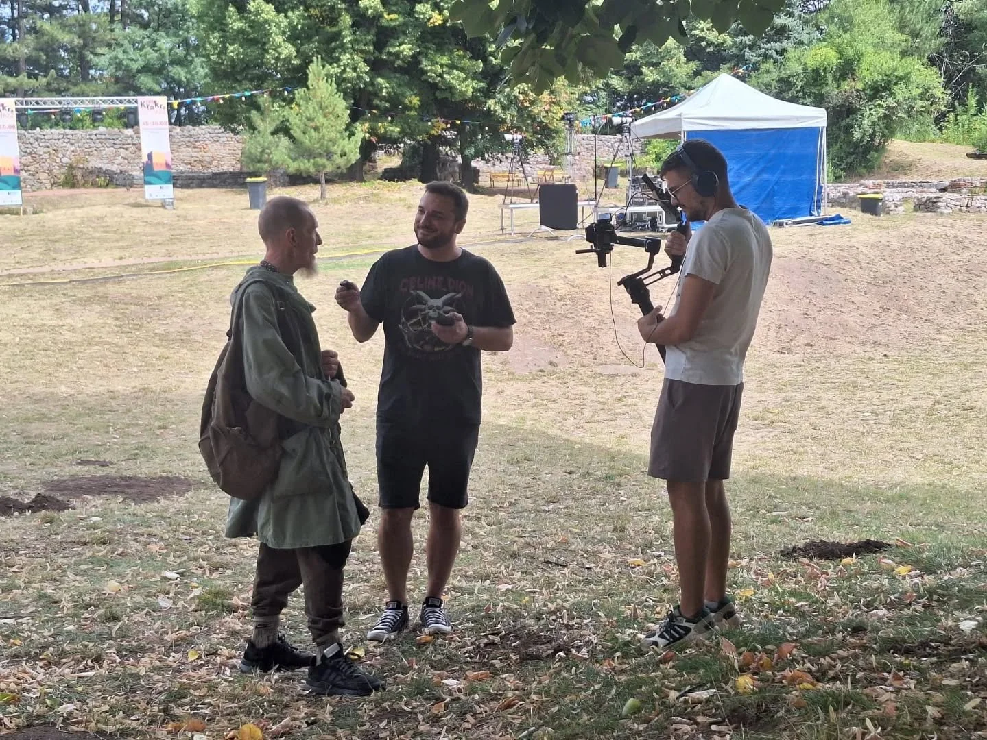 Three men are outdoors, engaged in conversation. One of them is holding a camera stabilizer, wearing headphones. The background features a blue tent, trees, and festival banners.