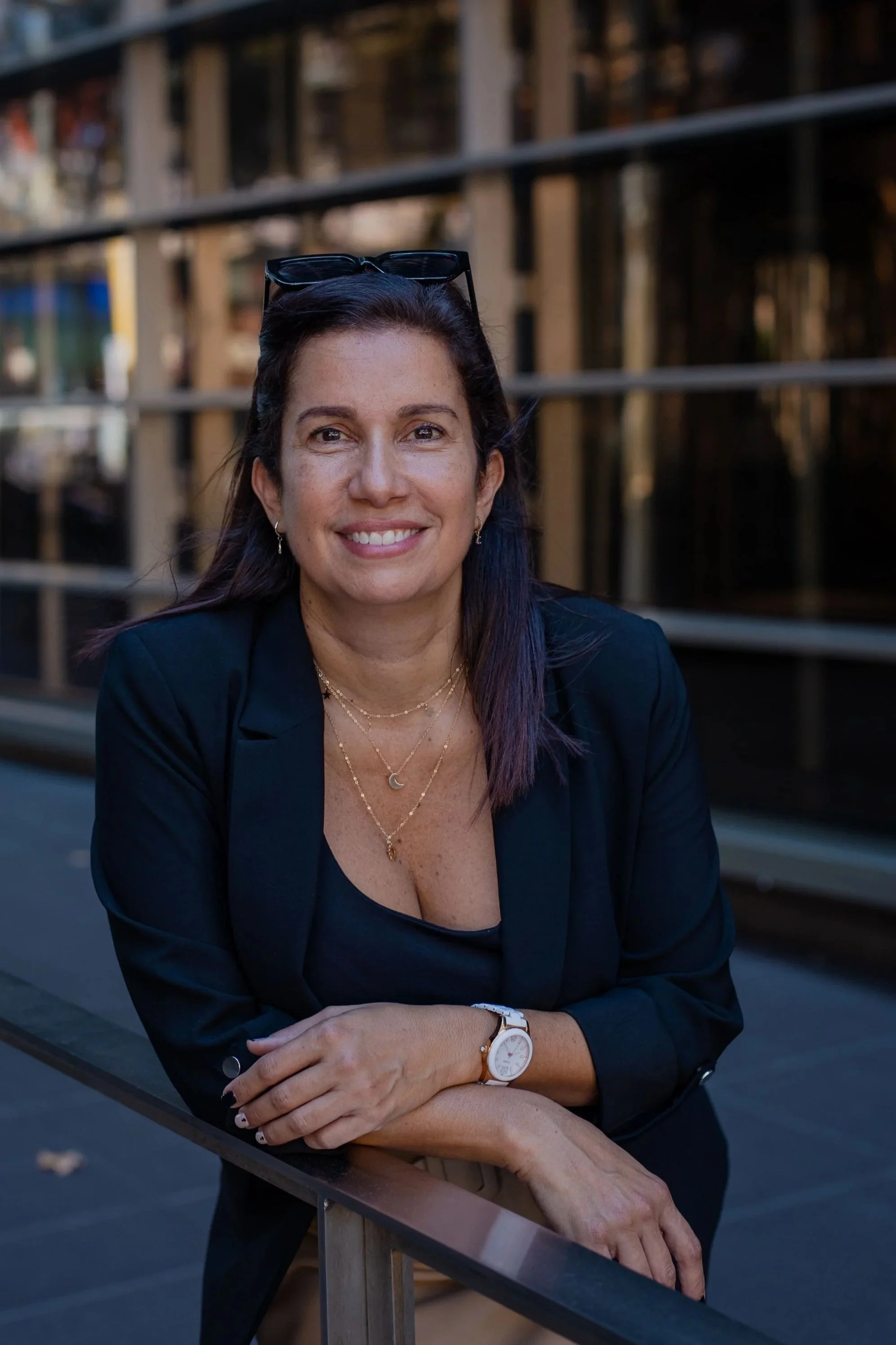 A woman with dark hair and glasses on her head, smiling, sitting outdoors against a city background with metal railings.