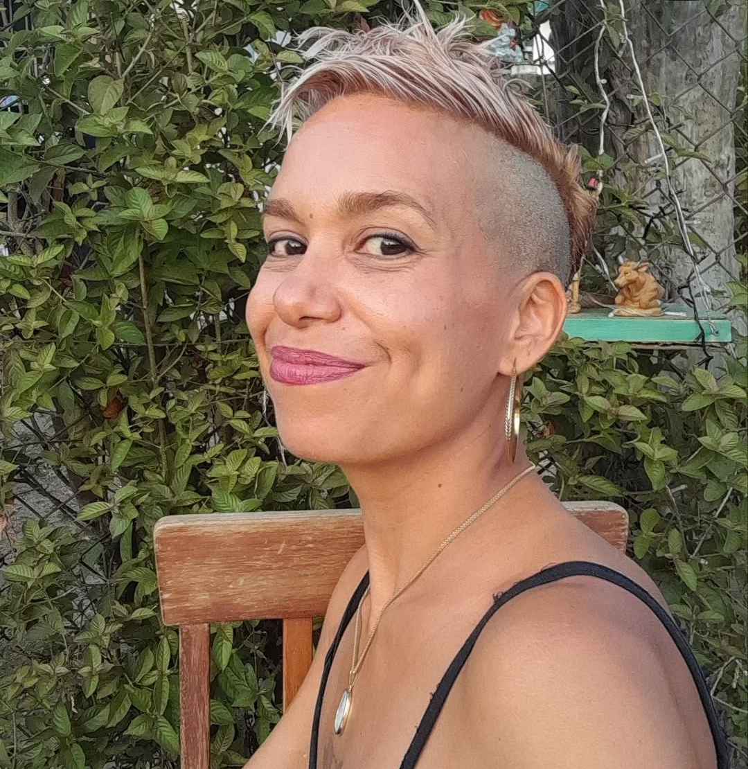 A woman with short, tousled blonde hair and an undercut, smiling and looking at the camera, sitting outdoors near green foliage with a wooden chair in the background.