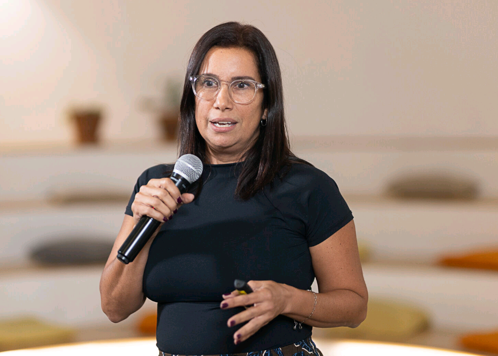 Mujer hablando en público con micrófono, en un lugar interior con fondo difuso.