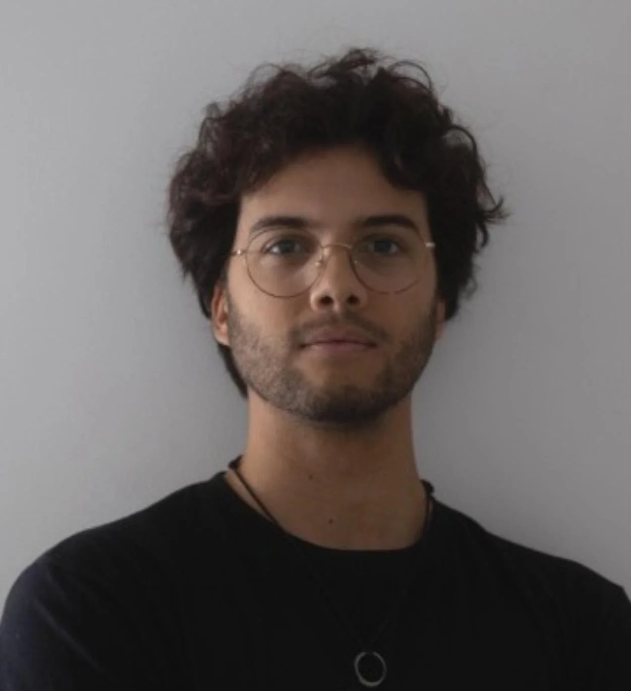 A man with curly dark hair, glasses, and a beard, wearing a black shirt and a necklace, standing against a plain gray wall.