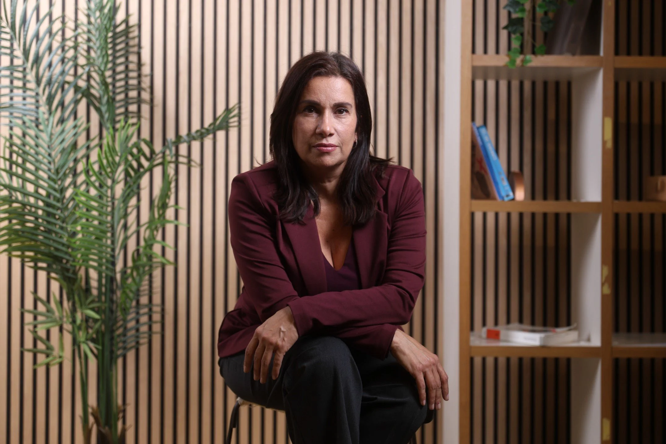 Mujer sentada en una silla, con fondo de paredes de listones de madera y estantería con libros y plantas.