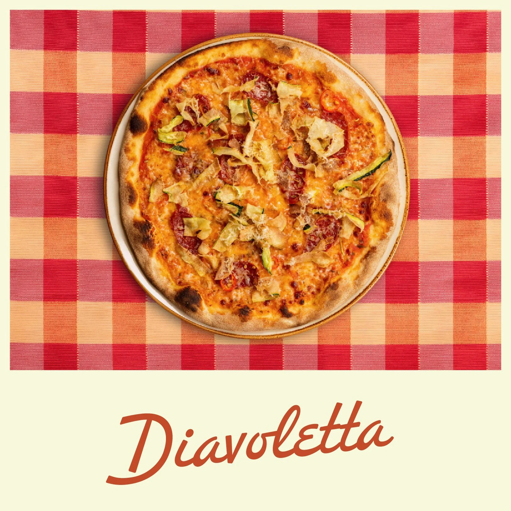 A pizza with toppings including bacon, zucchini, and cheese on a plate on a red and beige checkered tablecloth.