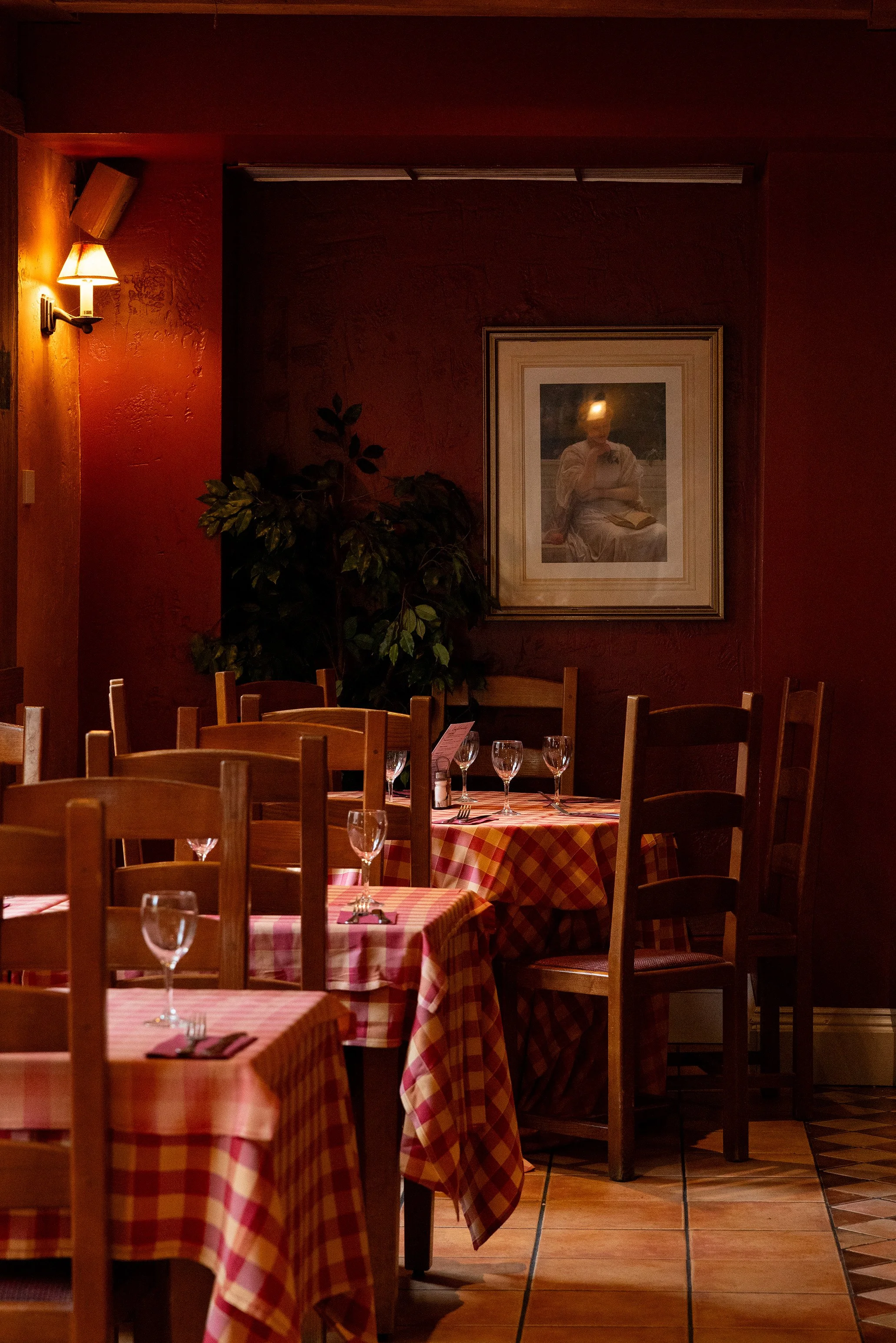 An empty restaurant dining area with wooden chairs and tables covered in red and white checkered tablecloths, set with wine glasses and silverware, dimly lit by wall-mounted lamps, with framed artwork on dark red walls and a potted plant in the corner.