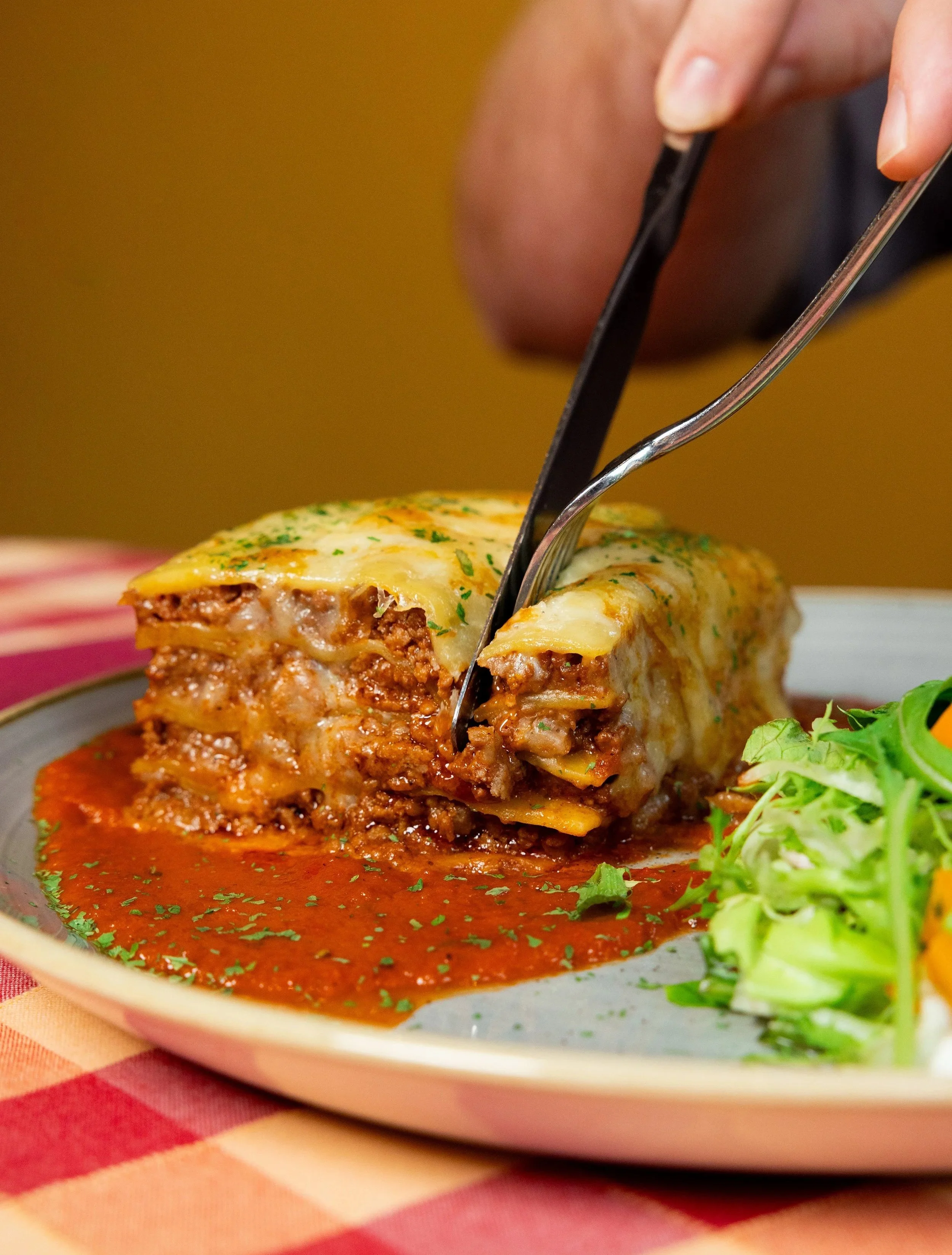 A close-up of a plate of lasagna with a side garden salad, with a person cutting a slice of lasagna with a fork and knife.